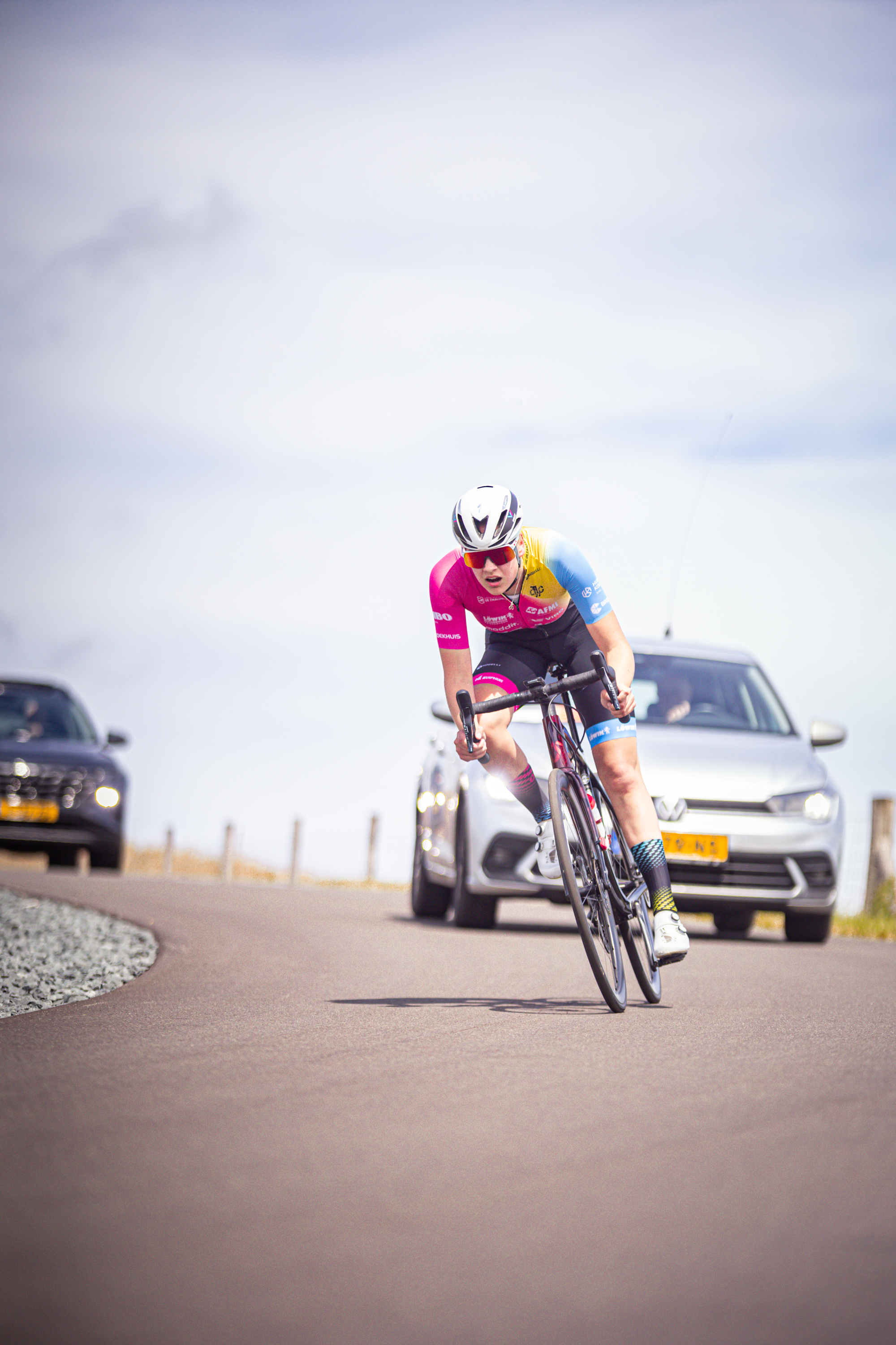 A cyclist wearing a pink outfit and white helmet is riding down the road past parked cars.