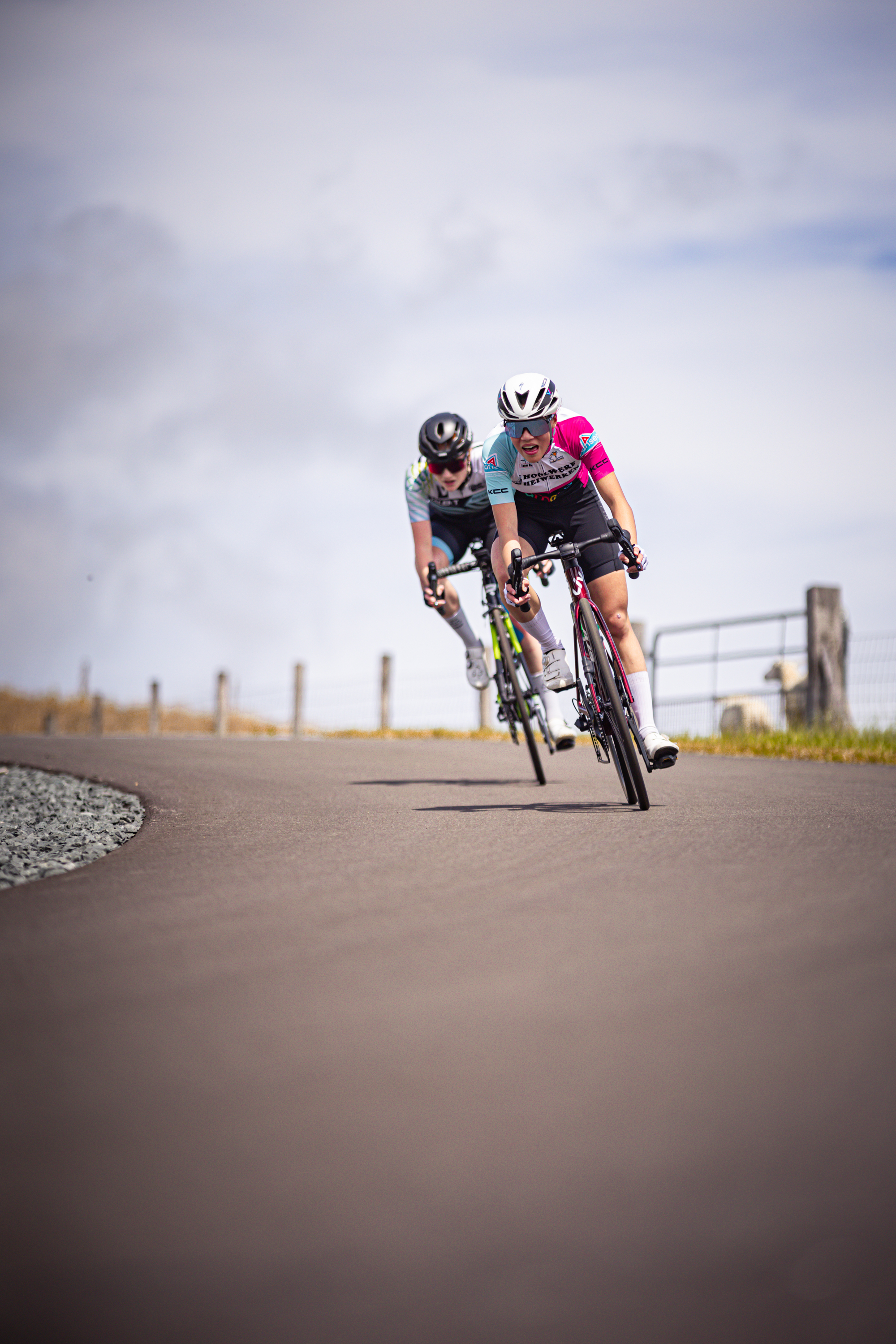 Two people are riding their bikes down a road, one is wearing a pink jersey and the other has a blue helmet on.