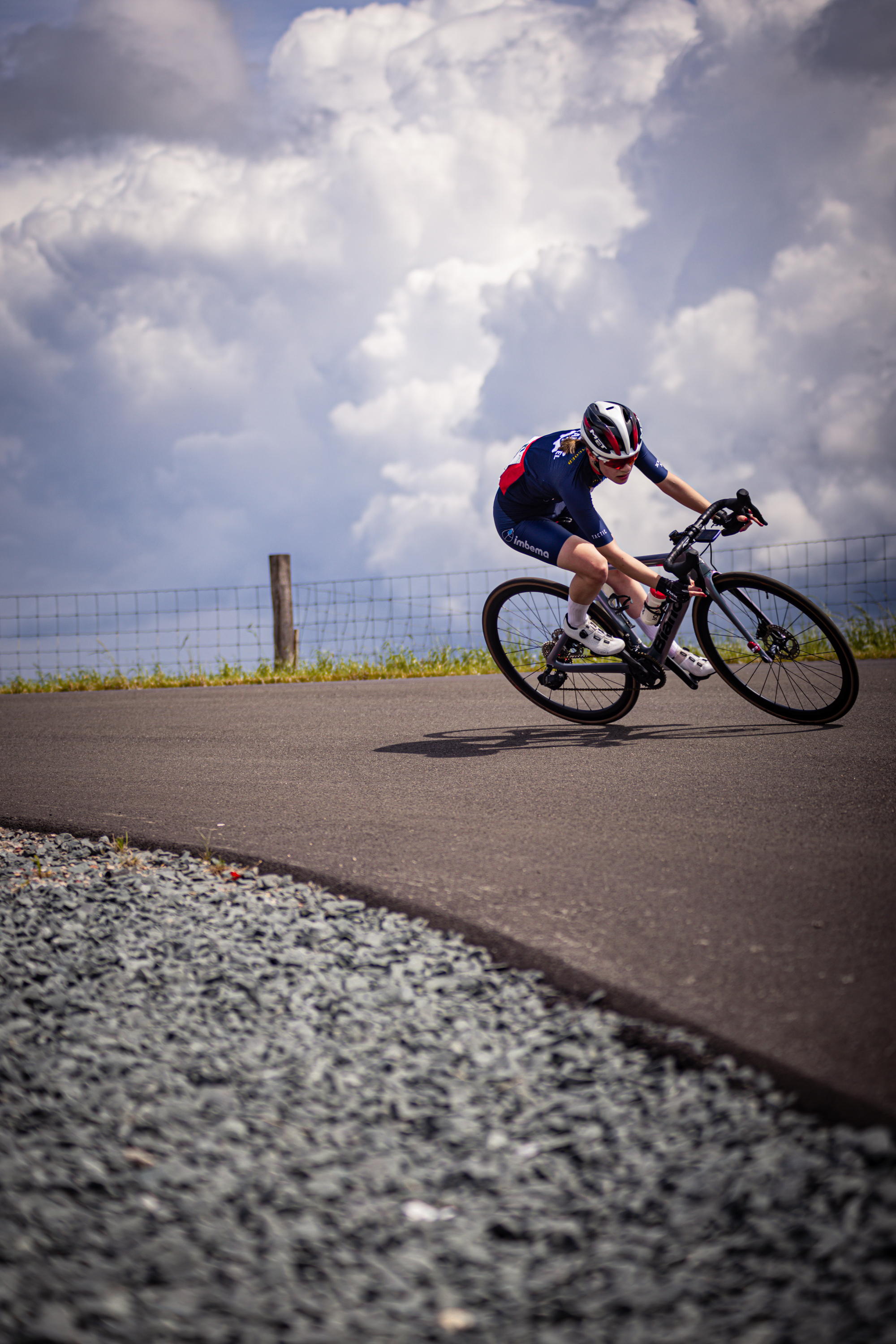 A man is riding his bike on a paved road. He has a blue and red jersey and a white helmet. The road is lined with gravel.