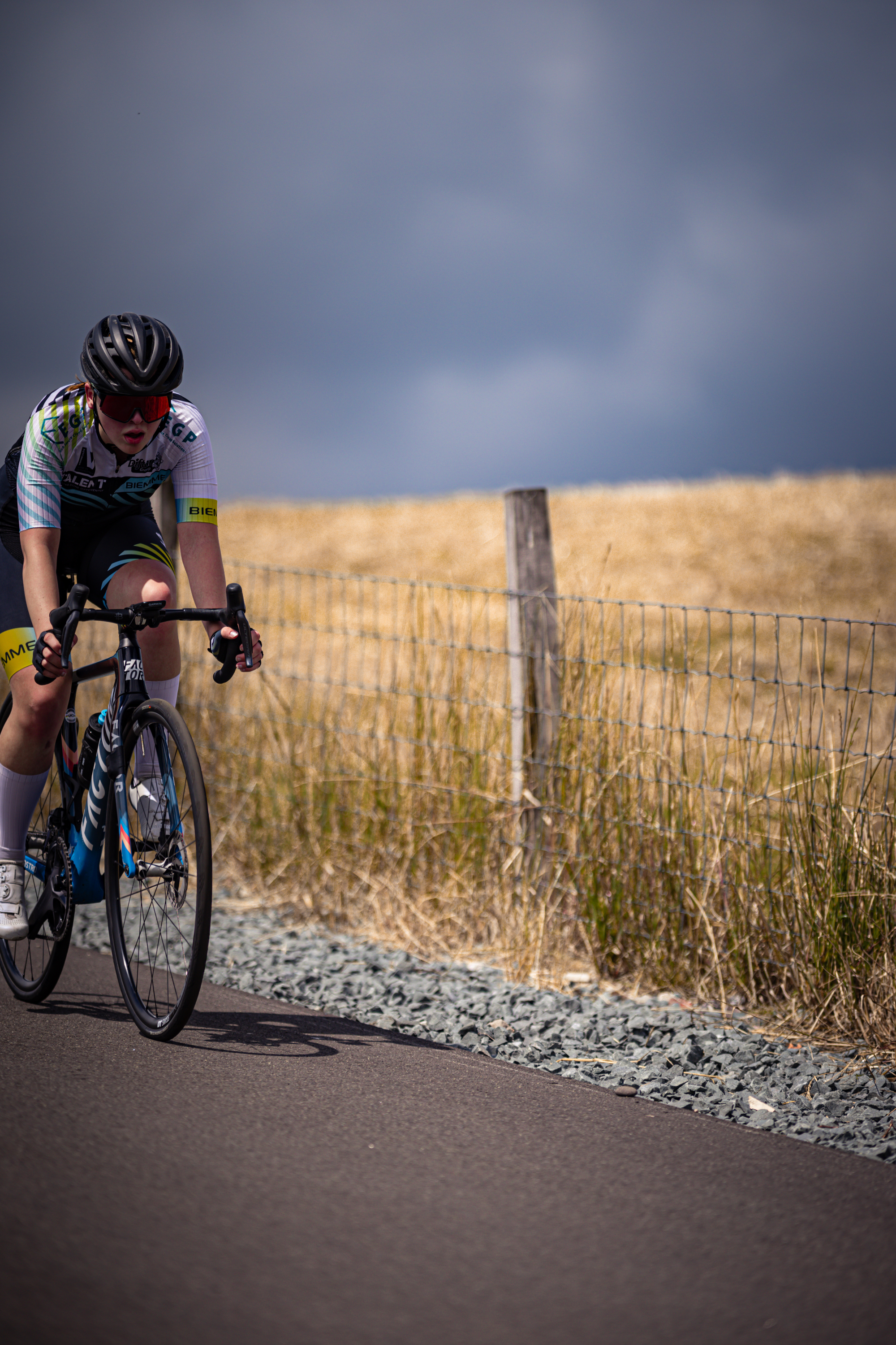 A person in a black and blue jersey rides a bike near a wooden fence.