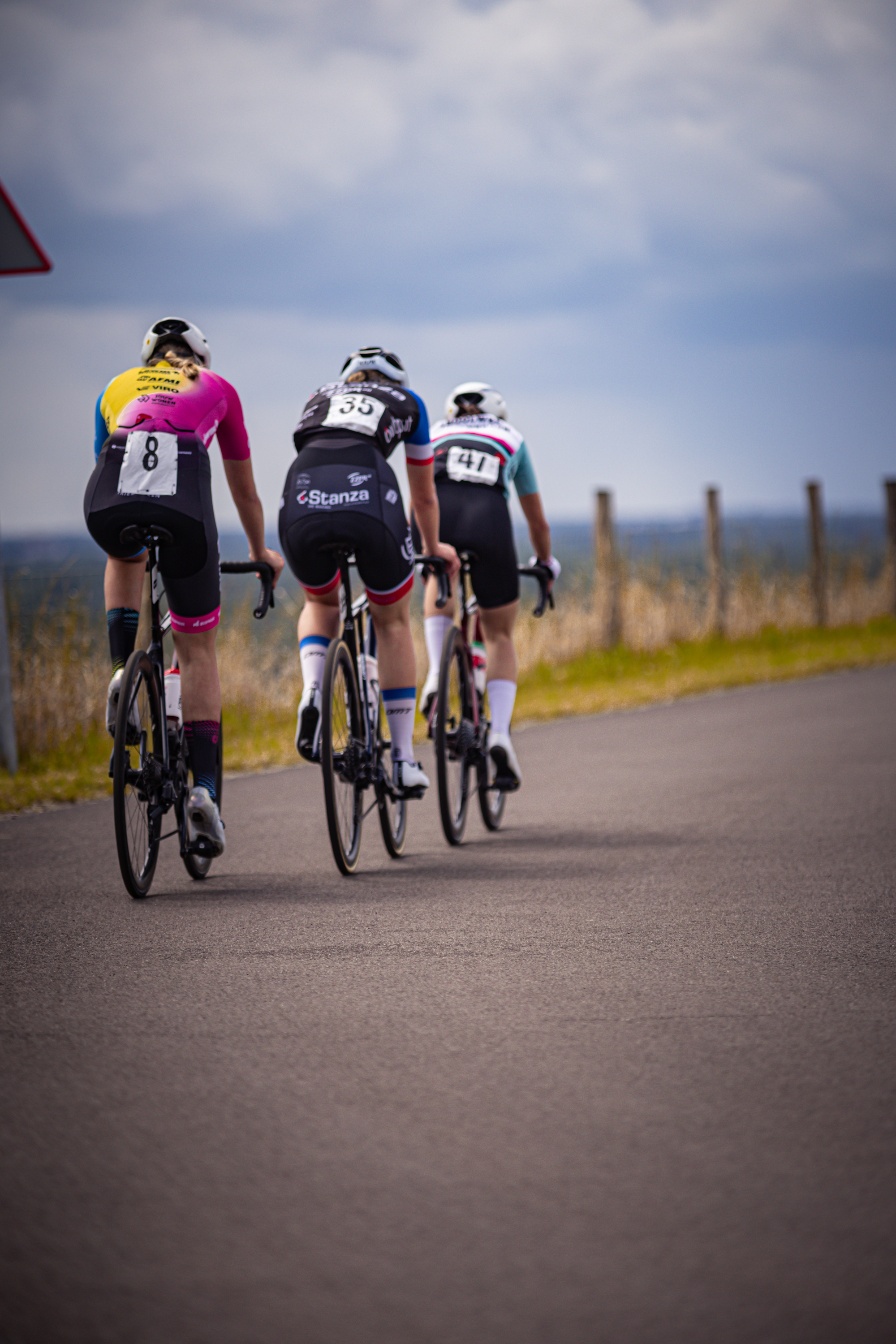 Three cyclists riding on the road in front of a fence.