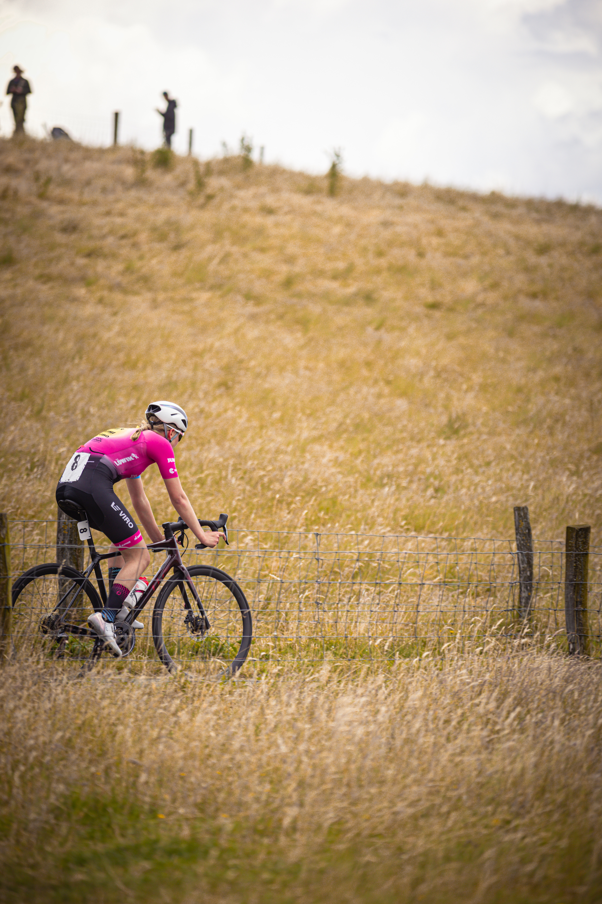 A cyclist in a pink jersey is riding down a hill near a fence.