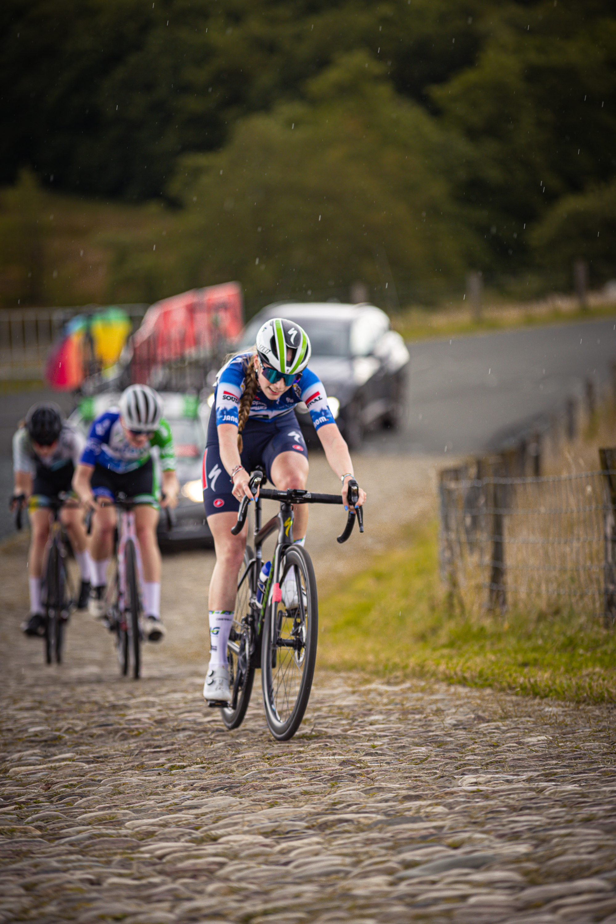 A girl in a green and blue uniform rides her bike through the cobblestone road, while two other girls chase behind.