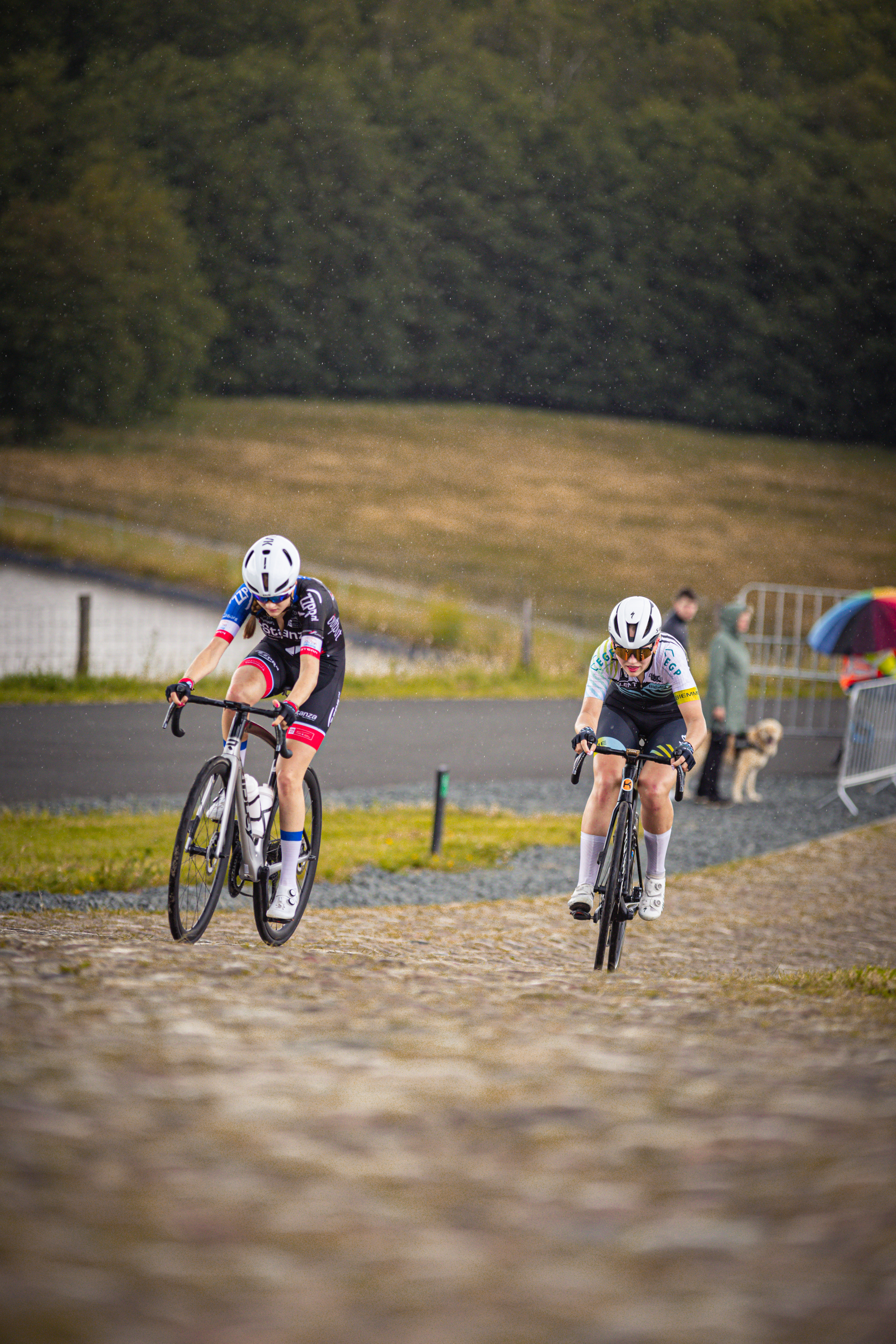 Two female cyclists race on a cobblestone road at the Nederlands Kampioenschap.