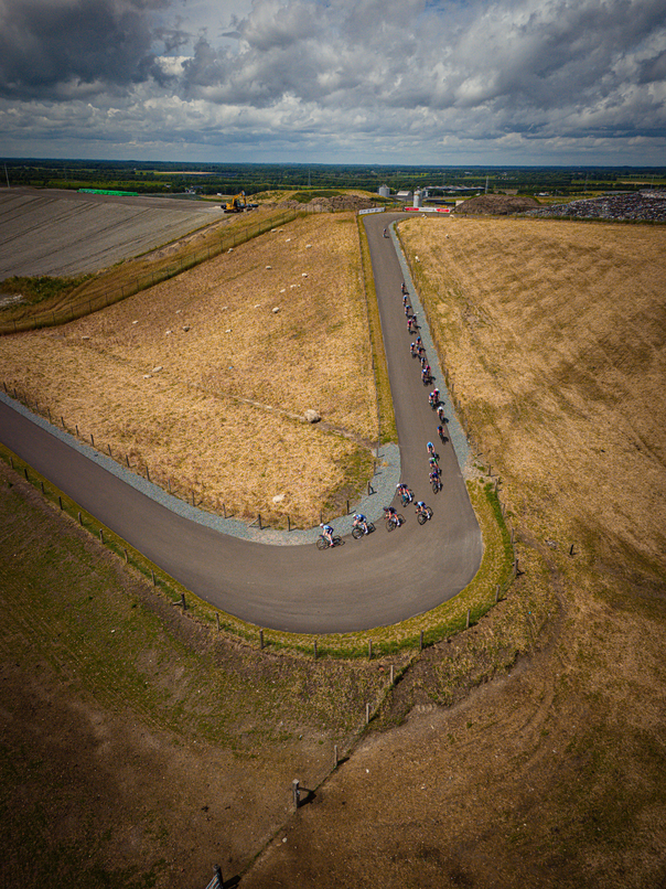 A group of cyclists riding a long, curved road. They are wearing numbers and helmets for safety. The sky is cloudy overhead.