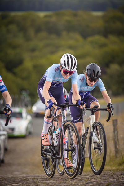 Two women in blue and white racing suits are riding bicycles during the Nederlands Kampioenschap race.