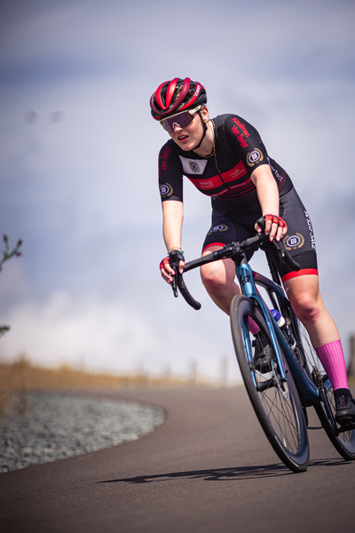 a woman wearing a red helmet and a black bicycle uniform riding her blue and white bike down a road.