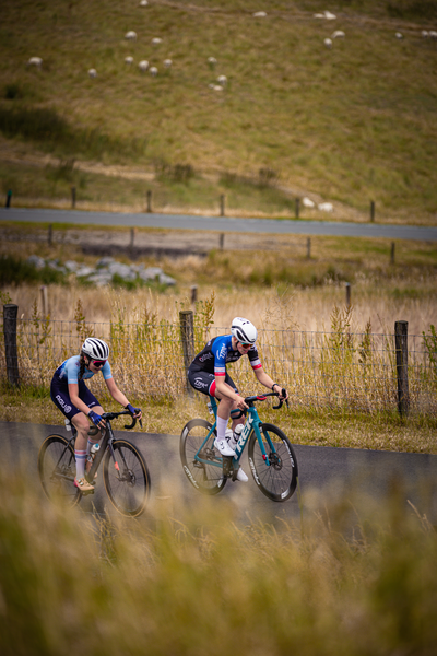 Two women are riding bicycles down a road. One of them is wearing a blue jersey with the number 23 on it.