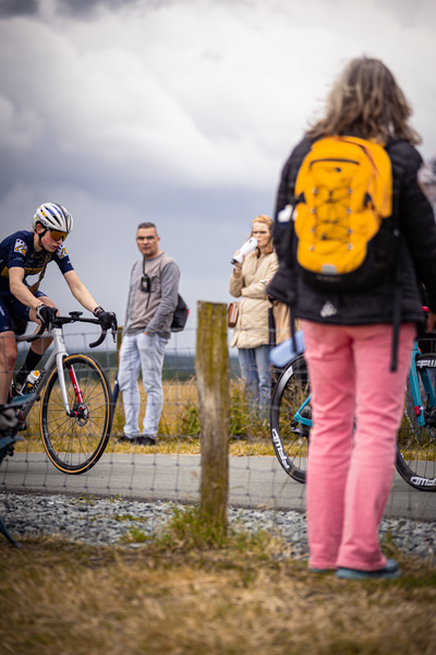 A group of cyclists stand around a fence, with one person wearing a yellow backpack.