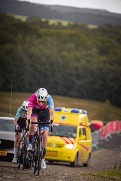 Two cyclists in the foreground of a race, with a yellow car behind them. They are wearing pink and black jerseys.