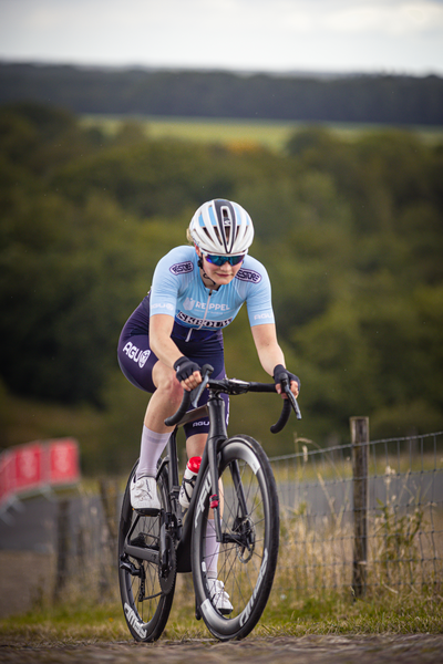 A woman wearing a blue and white jersey is riding a black bike. The word "Wielrennen" is visible on the jersey.