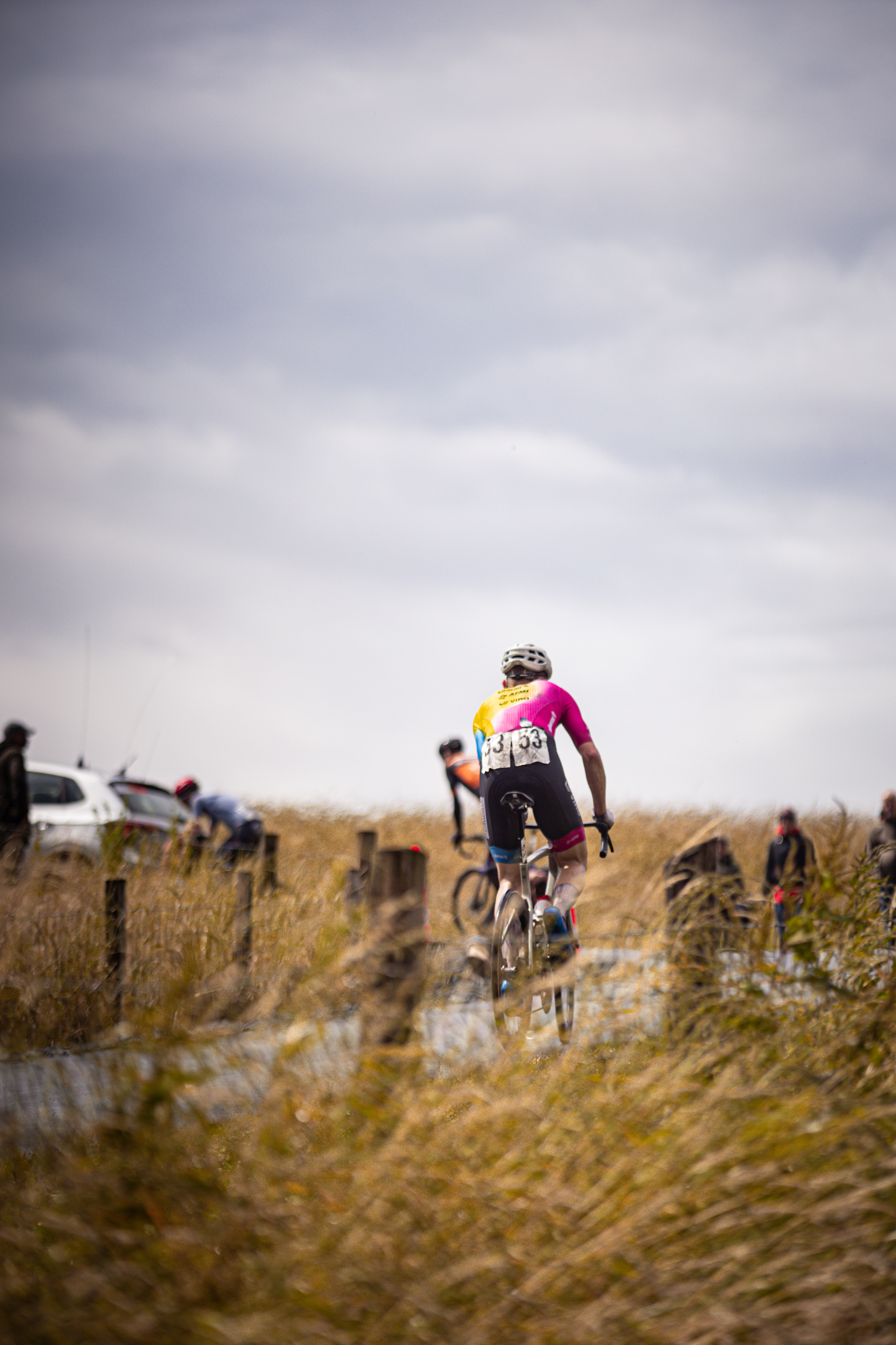 A cyclist wearing a pink shirt with the letters ZC on it is riding his bike on a dirt road near a field.