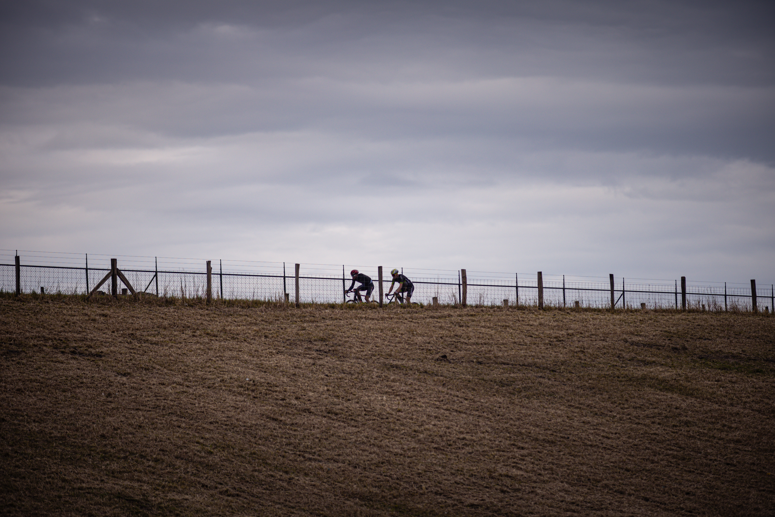 Several cyclists are riding on a dirt hill, with a barbed wire fence in front of them.