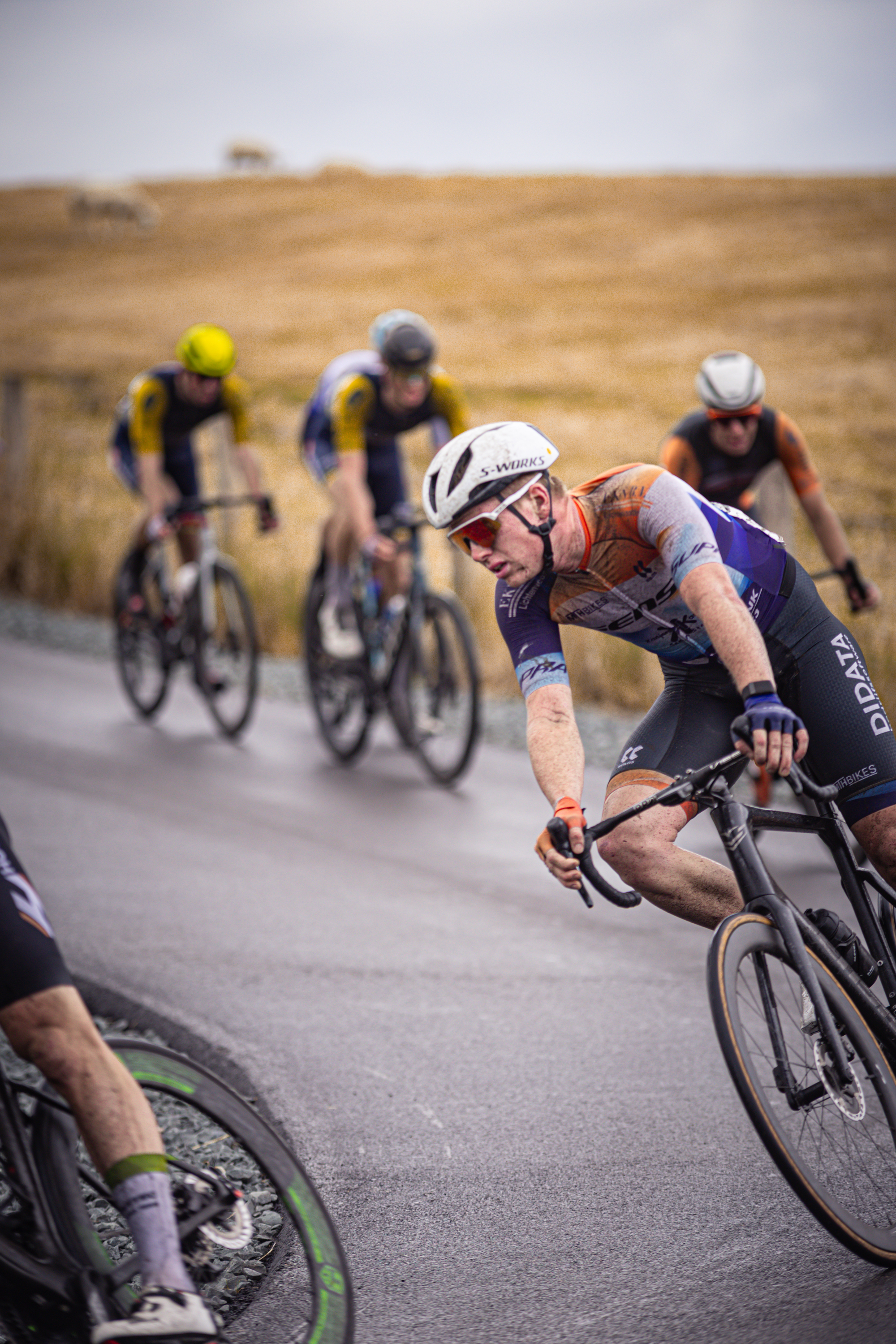 Five cyclists racing in a group, with one cyclist ahead of the others. The man leading has red hair and is wearing glasses.