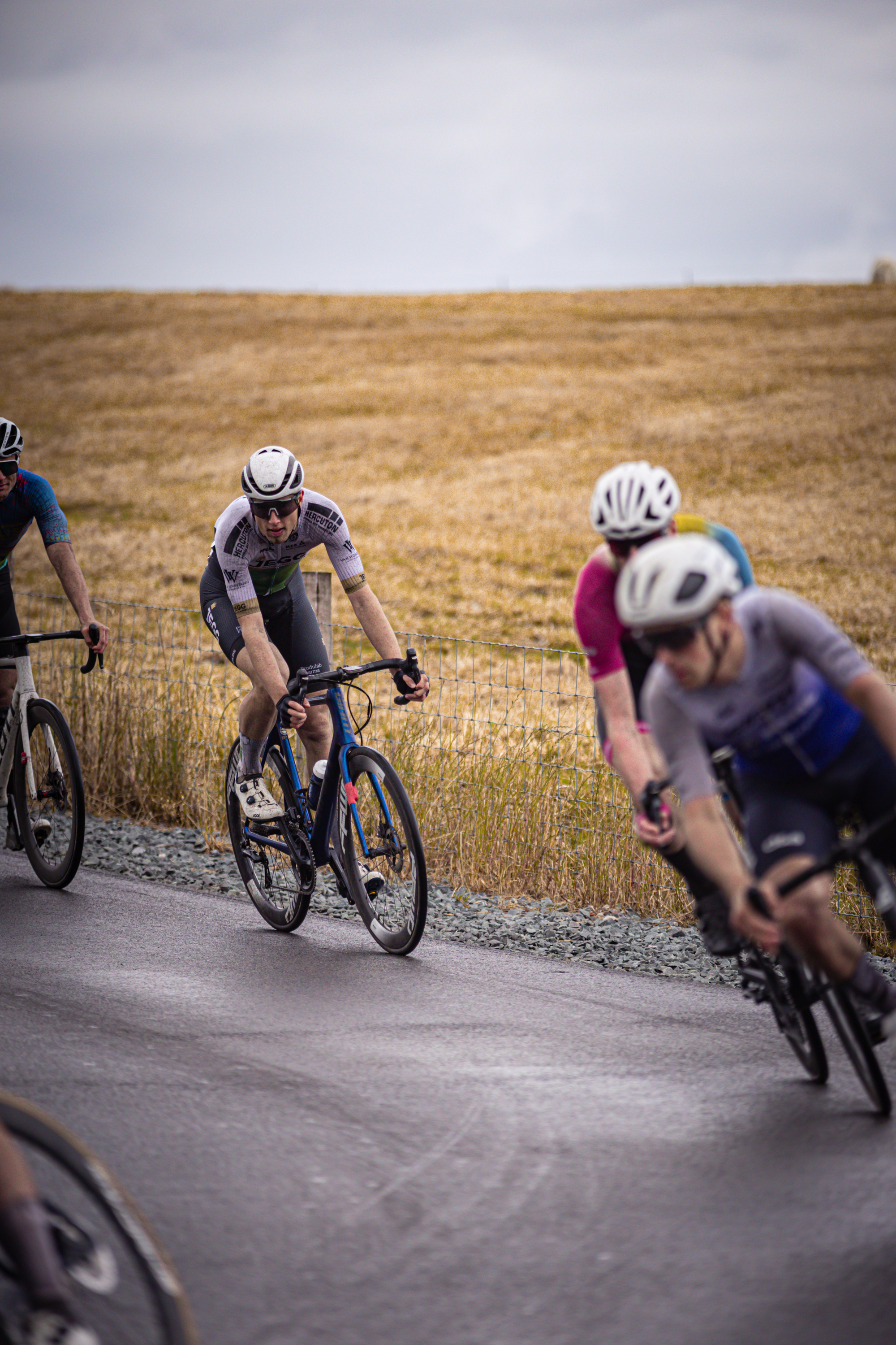 Four cyclists racing in a field on a cloudy day. They are all wearing helmets and are riding bikes.