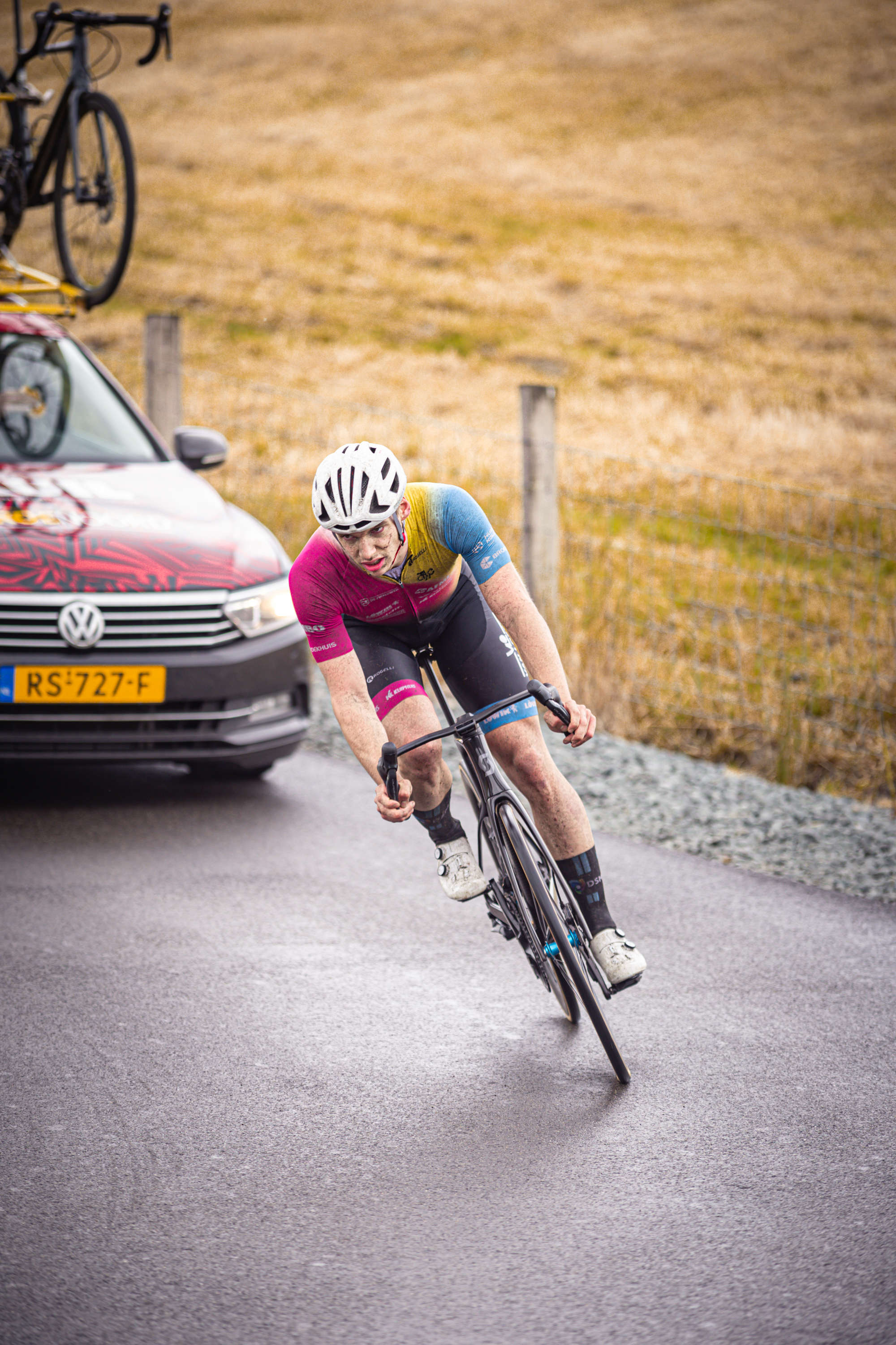 A person in a pink shirt riding a bike on the street with grass and a car in the background.