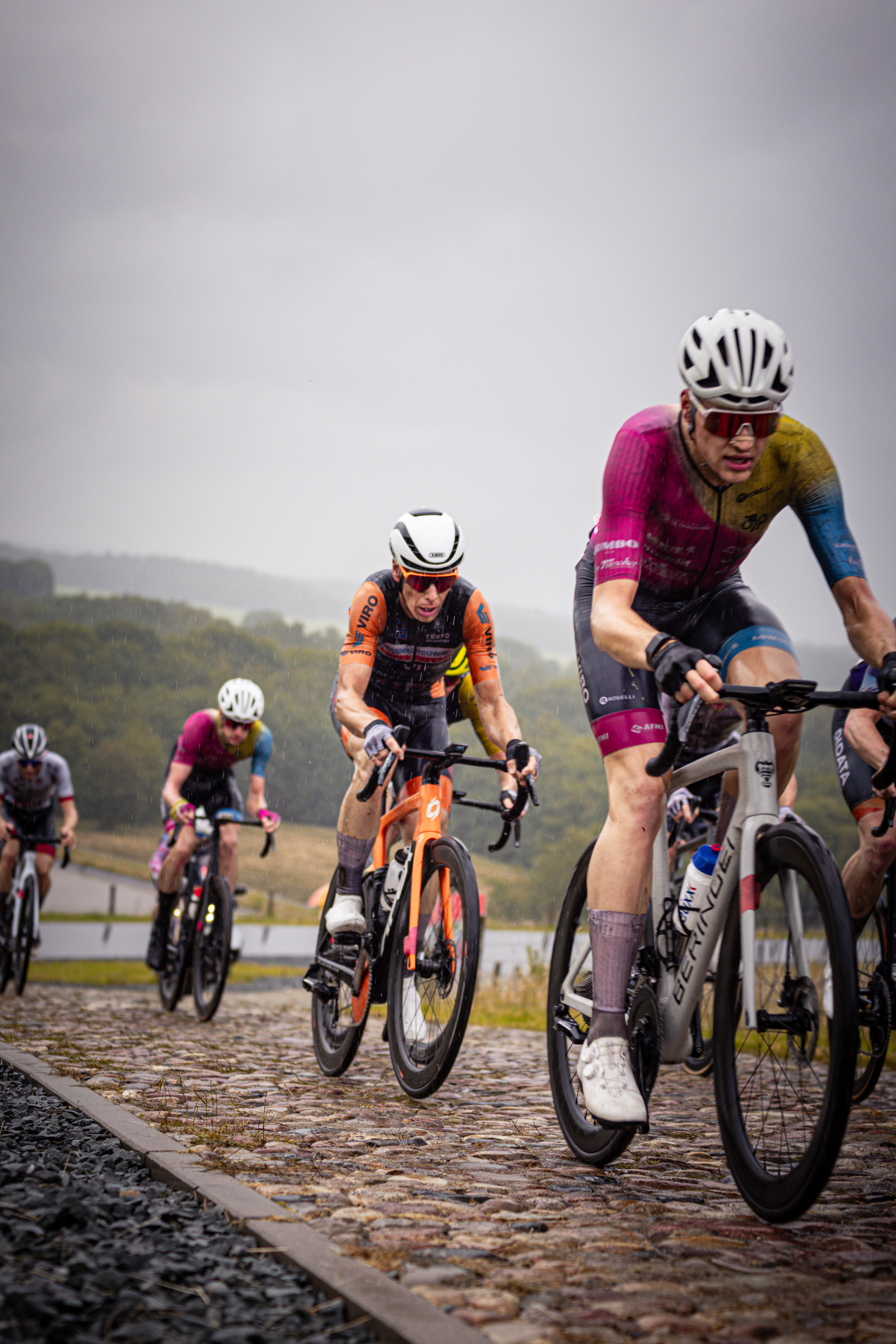 Wielrenners on a track with one wearing an orange shirt and the other two in blue shirts.