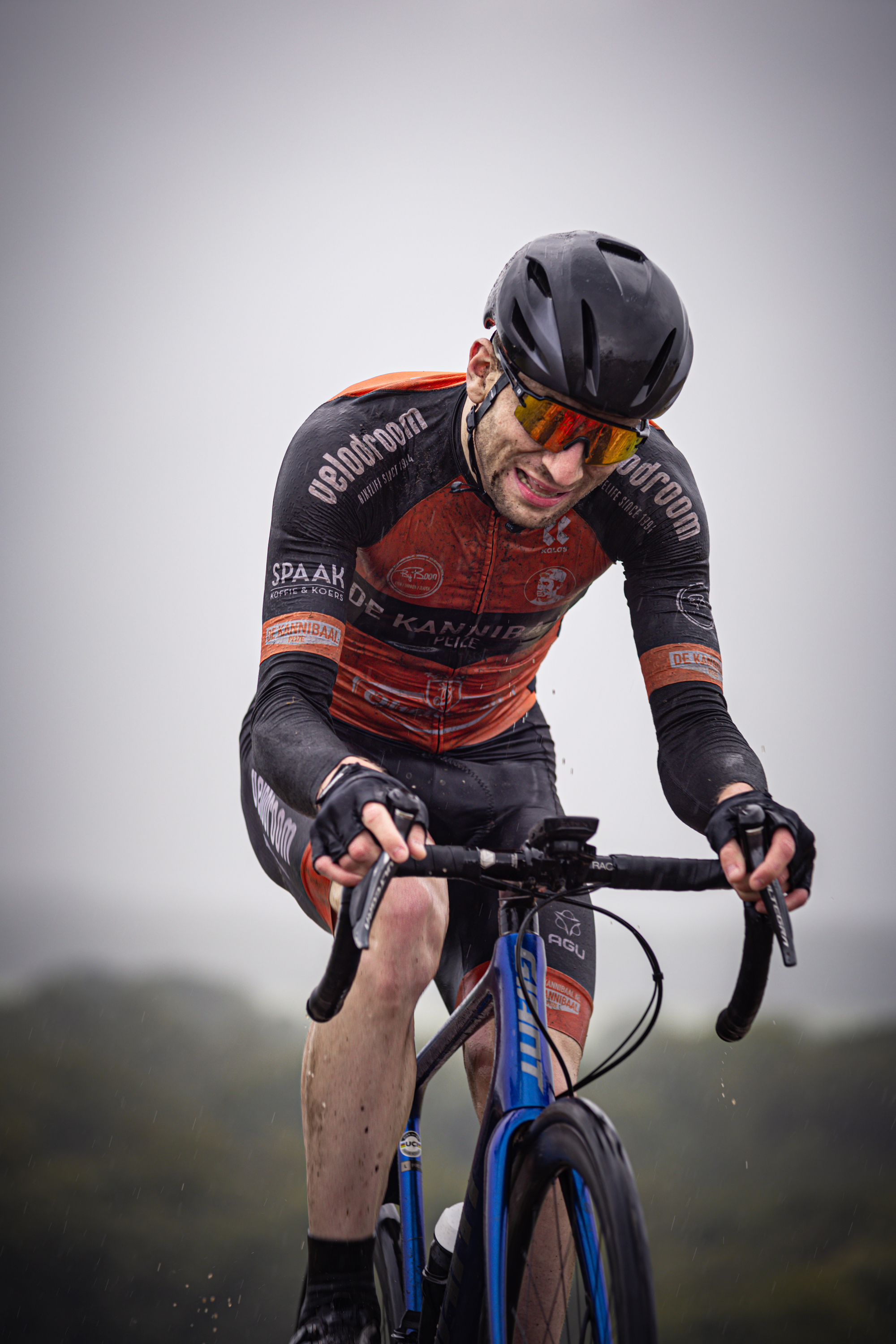 A man wearing a black and red bike suit riding a bicycle with the word "wielrennen" on his helmet.