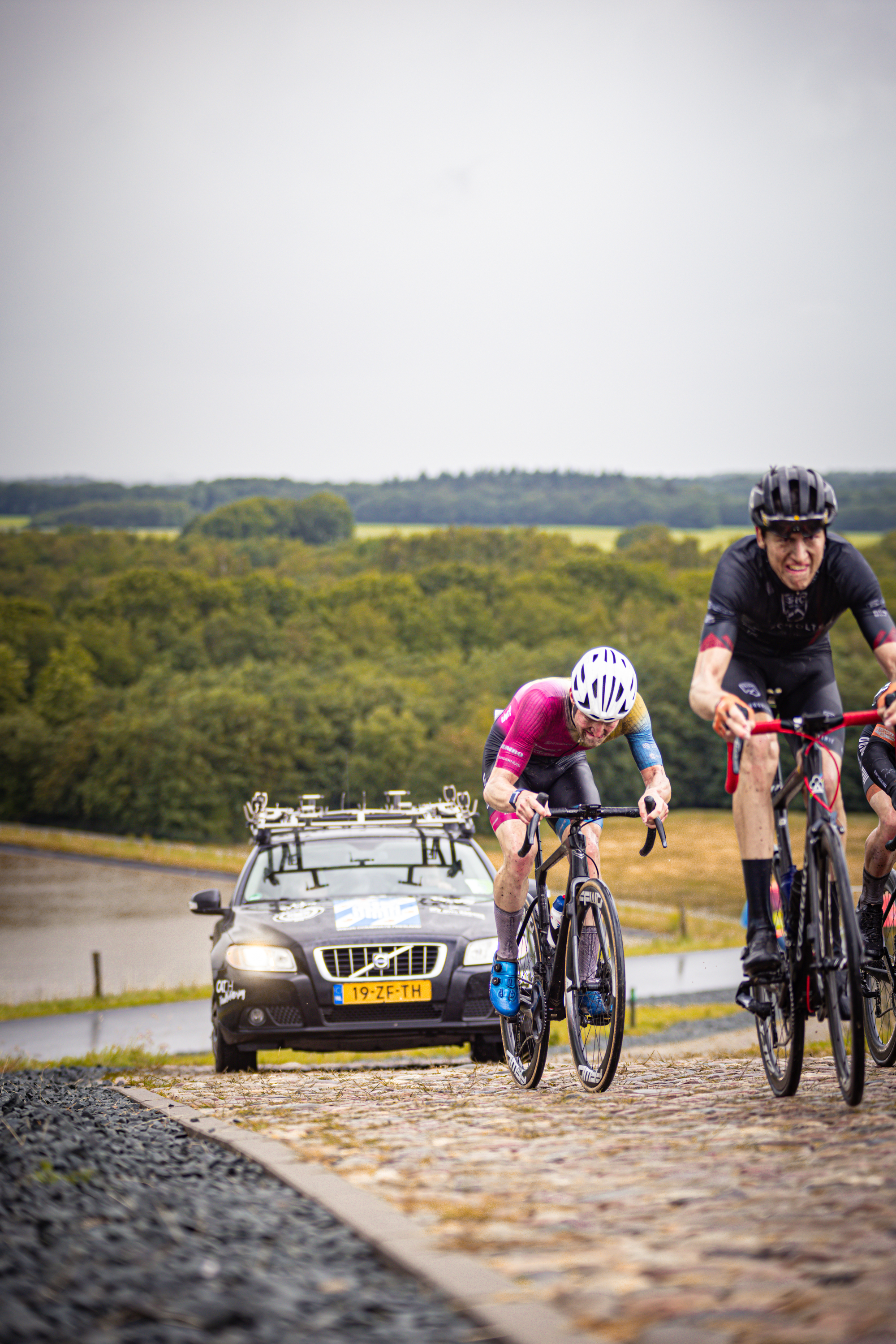 Two cyclists on a cobblestone road. The man in the black and red shirt has his mouth open.