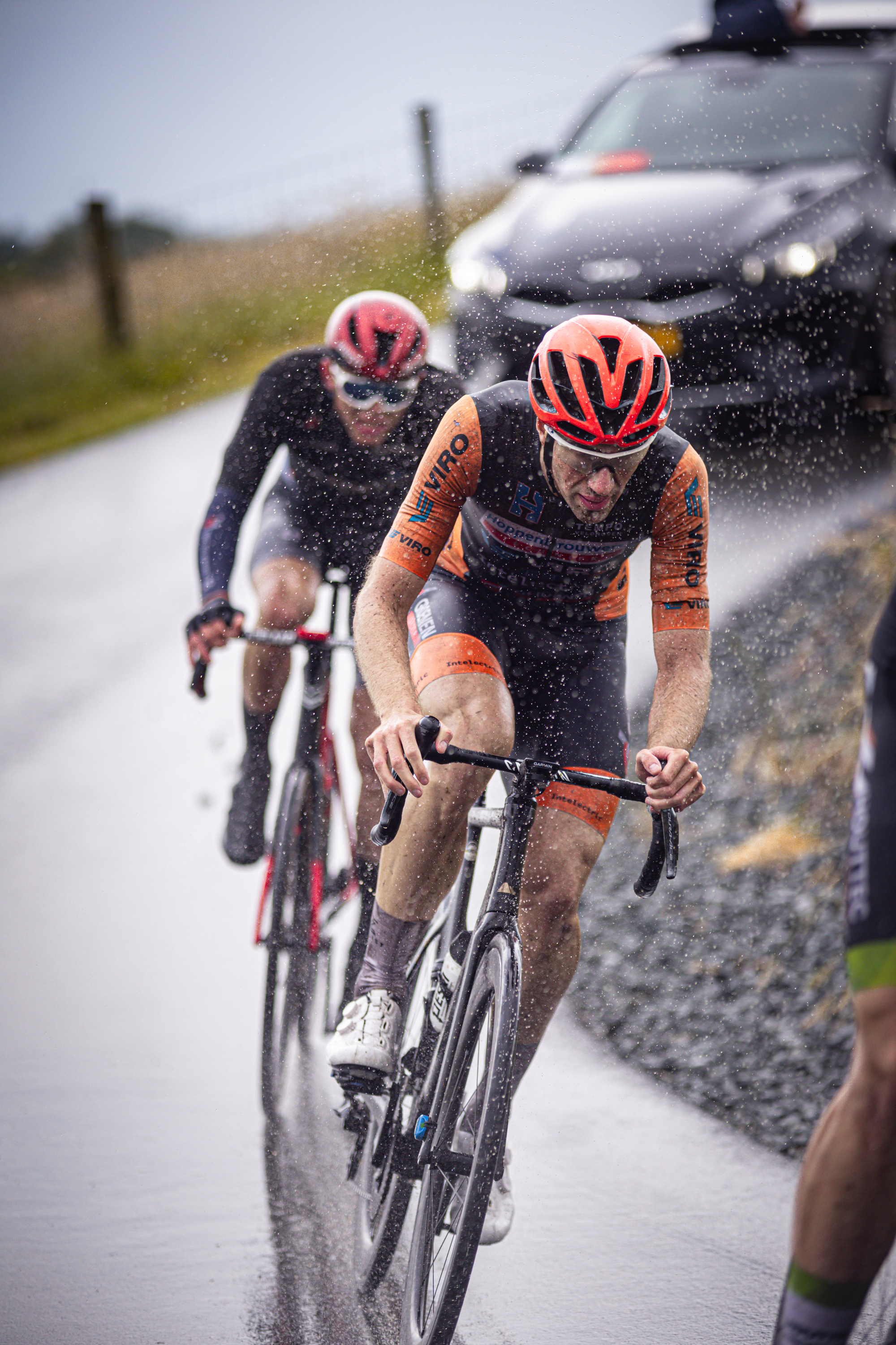 Two male cyclists wearing helmets are riding on a wet road.