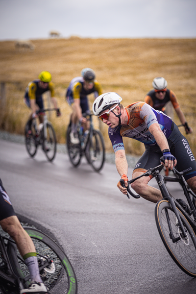 Five cyclists racing in a group, with one cyclist ahead of the others. The man leading has red hair and is wearing glasses.