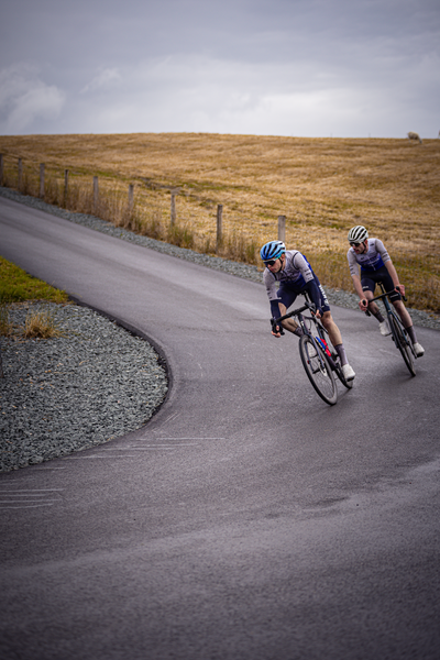 Two cyclists are riding on a paved road with a grassy hill in the background.