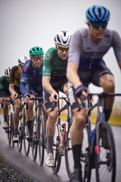 A group of cyclists are riding down a wet road, possibly in the Nederlands Kampioenschap.