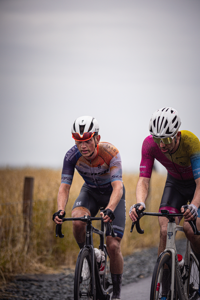 Two cyclists wearing helmets and colorful jerseys are racing on a gravel road.