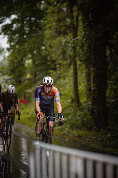 A cyclist wearing a blue jersey and helmet is riding on a wet road near trees.