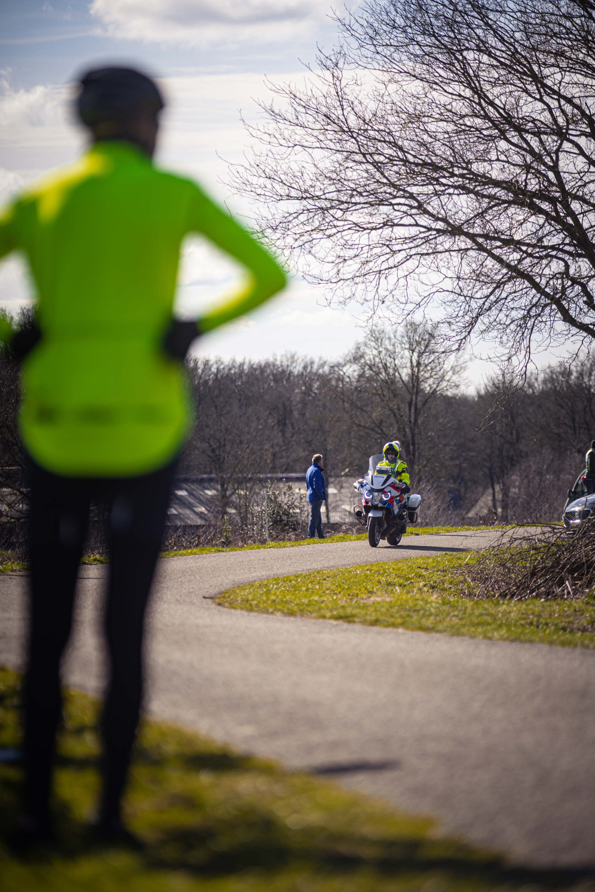 A person in a yellow jacket stands next to the road while two men on motorcycles drive down it.