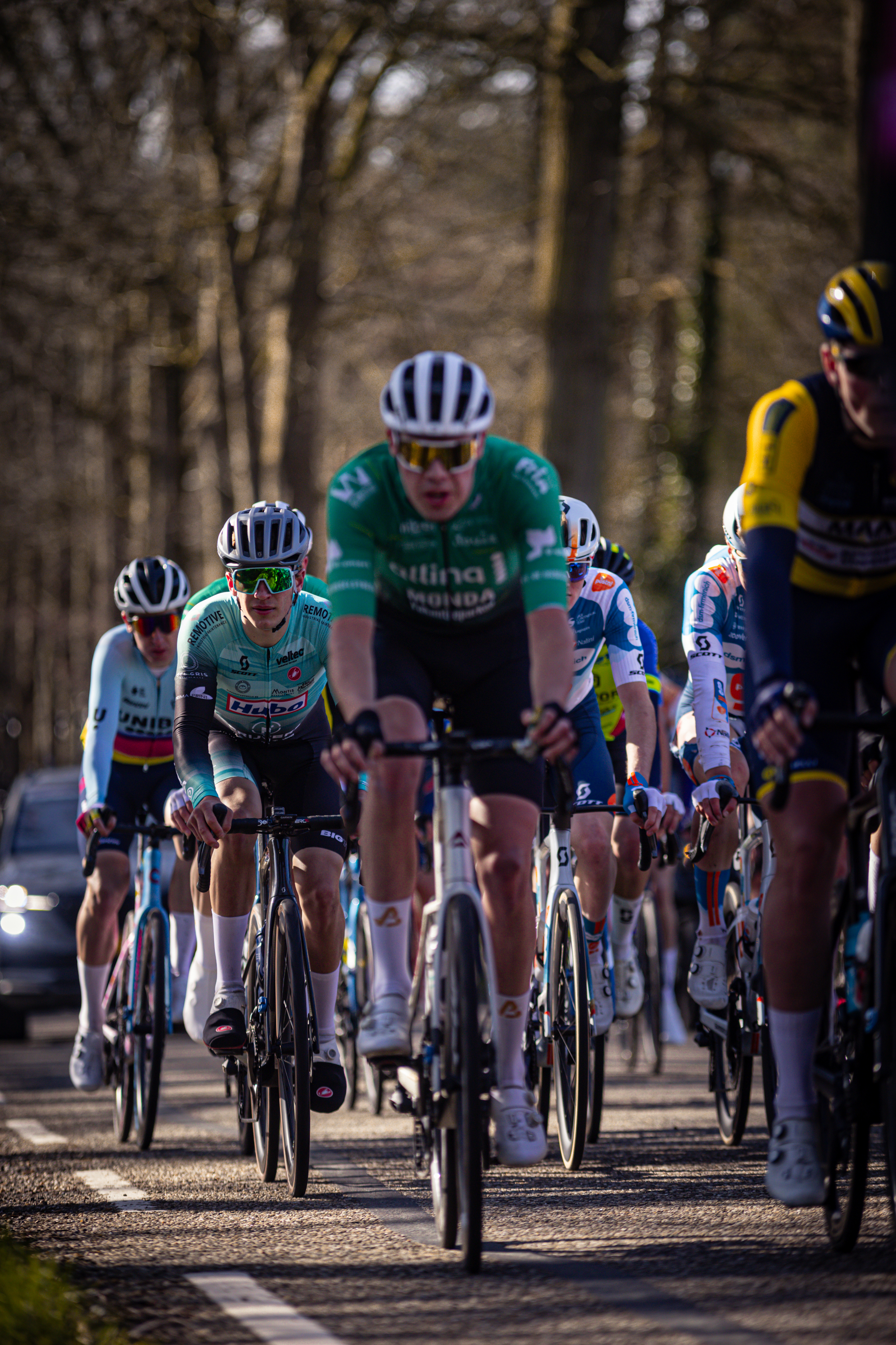 A group of cyclists wearing Wielrennen gear race down a road.