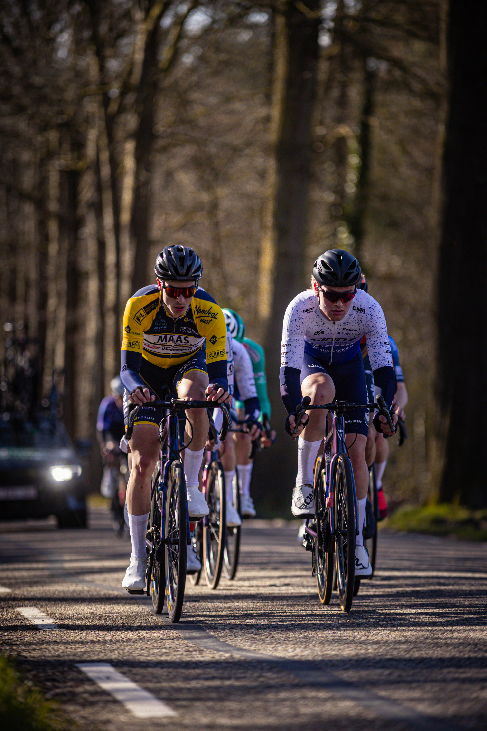 Two cyclists are wearing matching yellow and white uniforms while racing on a path near woods.