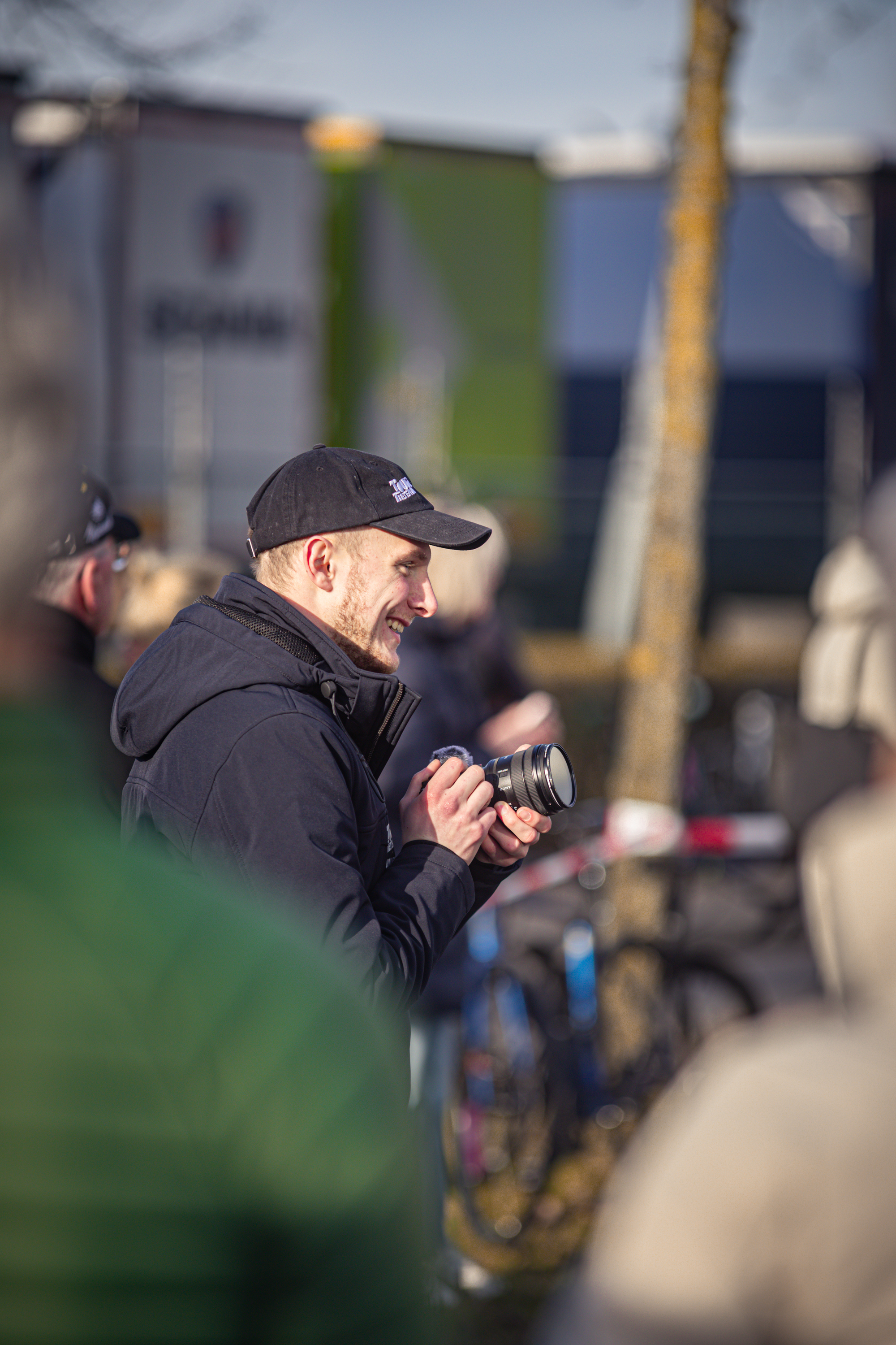 A man in a black jacket holds up his camera at the Wielrennen event.