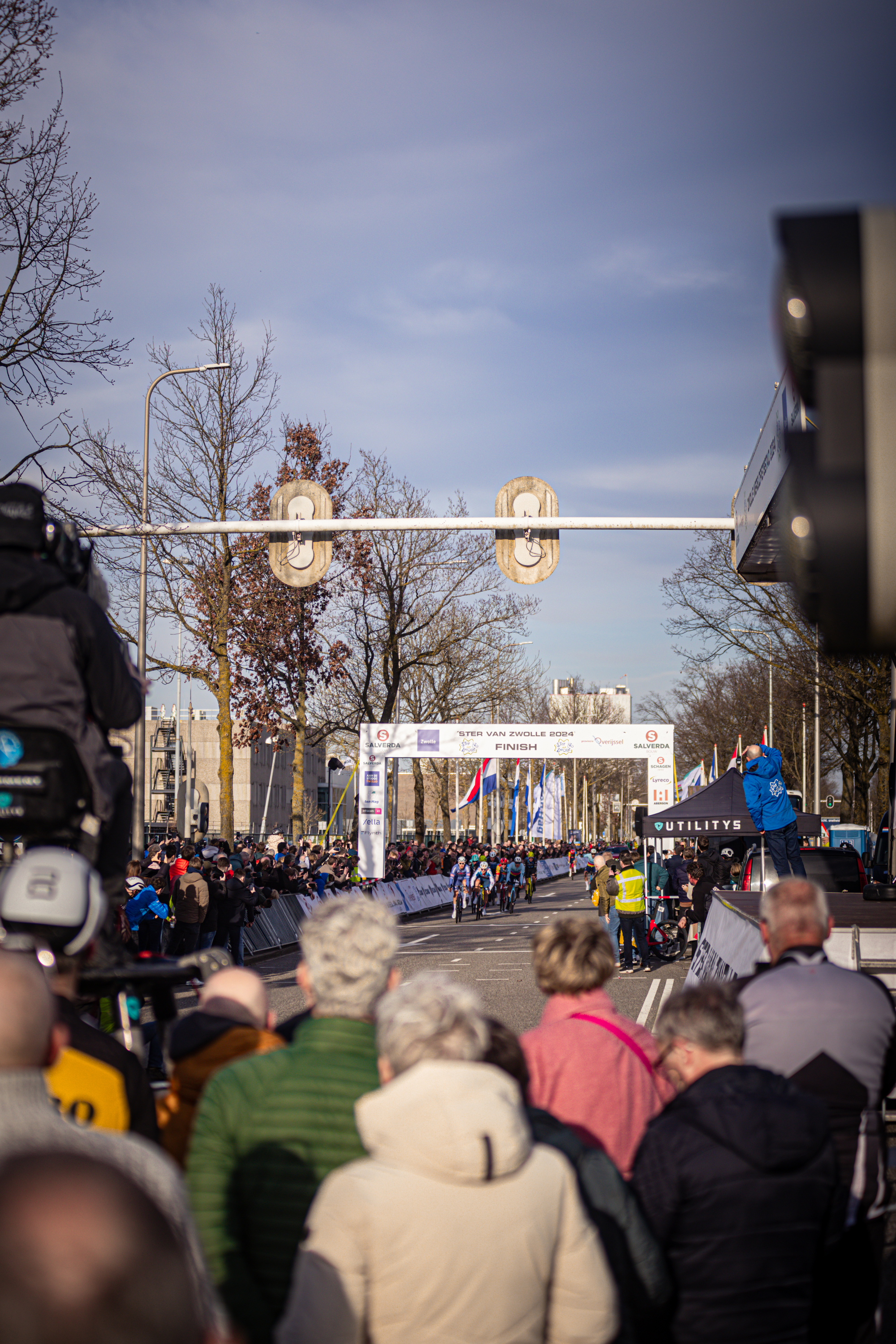 A group of people stand under a street sign for the Ster van Zwolle race. They are waiting for the cyclists to pass by.