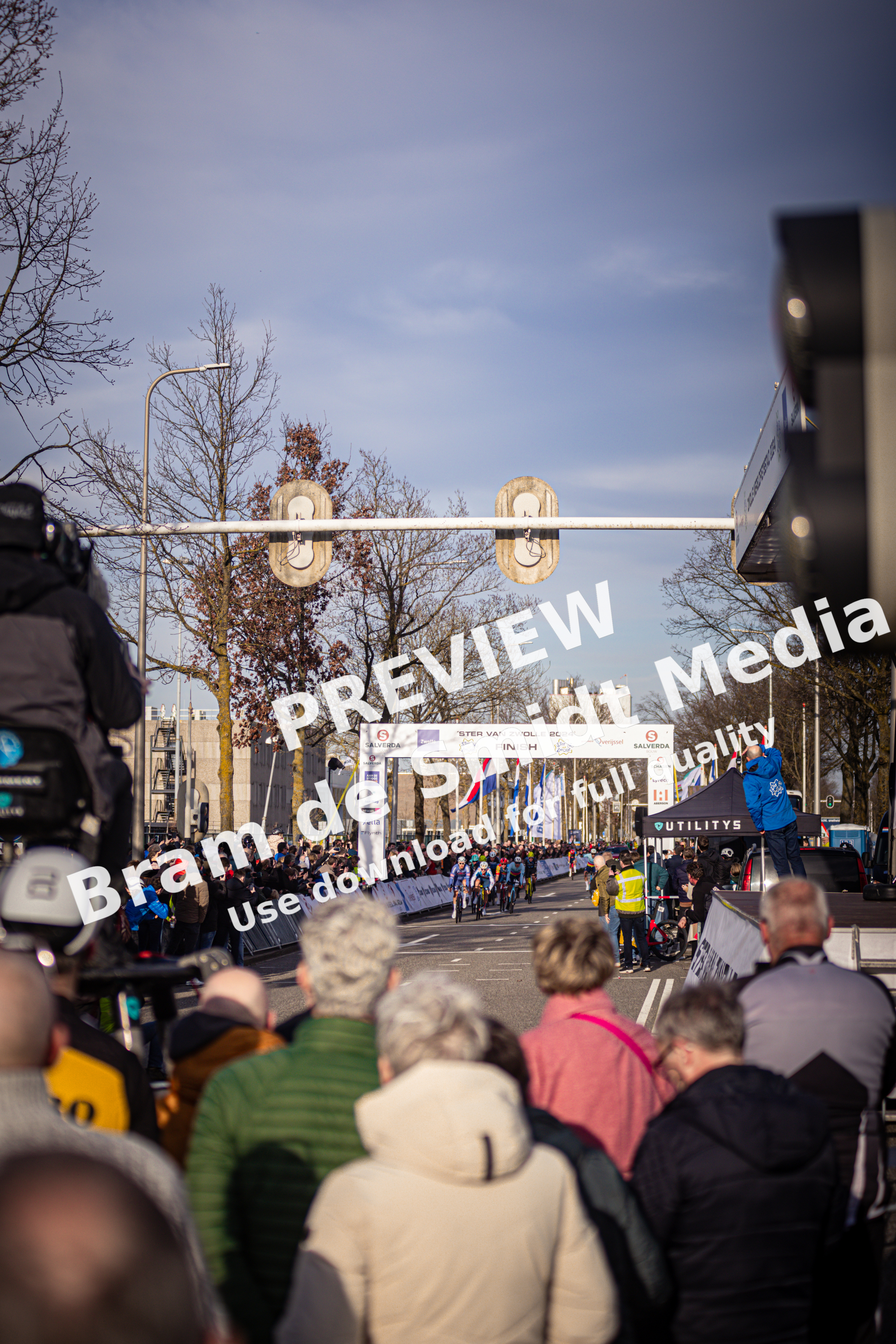 A group of people stand under a street sign for the Ster van Zwolle race. They are waiting for the cyclists to pass by.