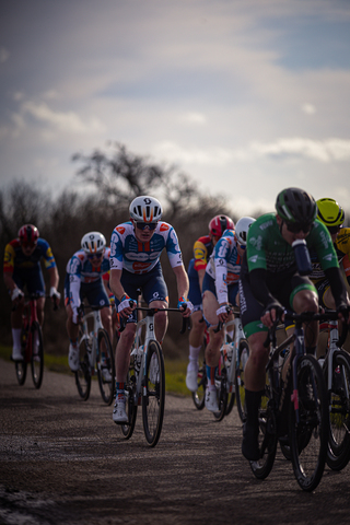 A group of cyclists race down a dirt road. One rider in the front wears a green jersey that says Ster van Zwolle.