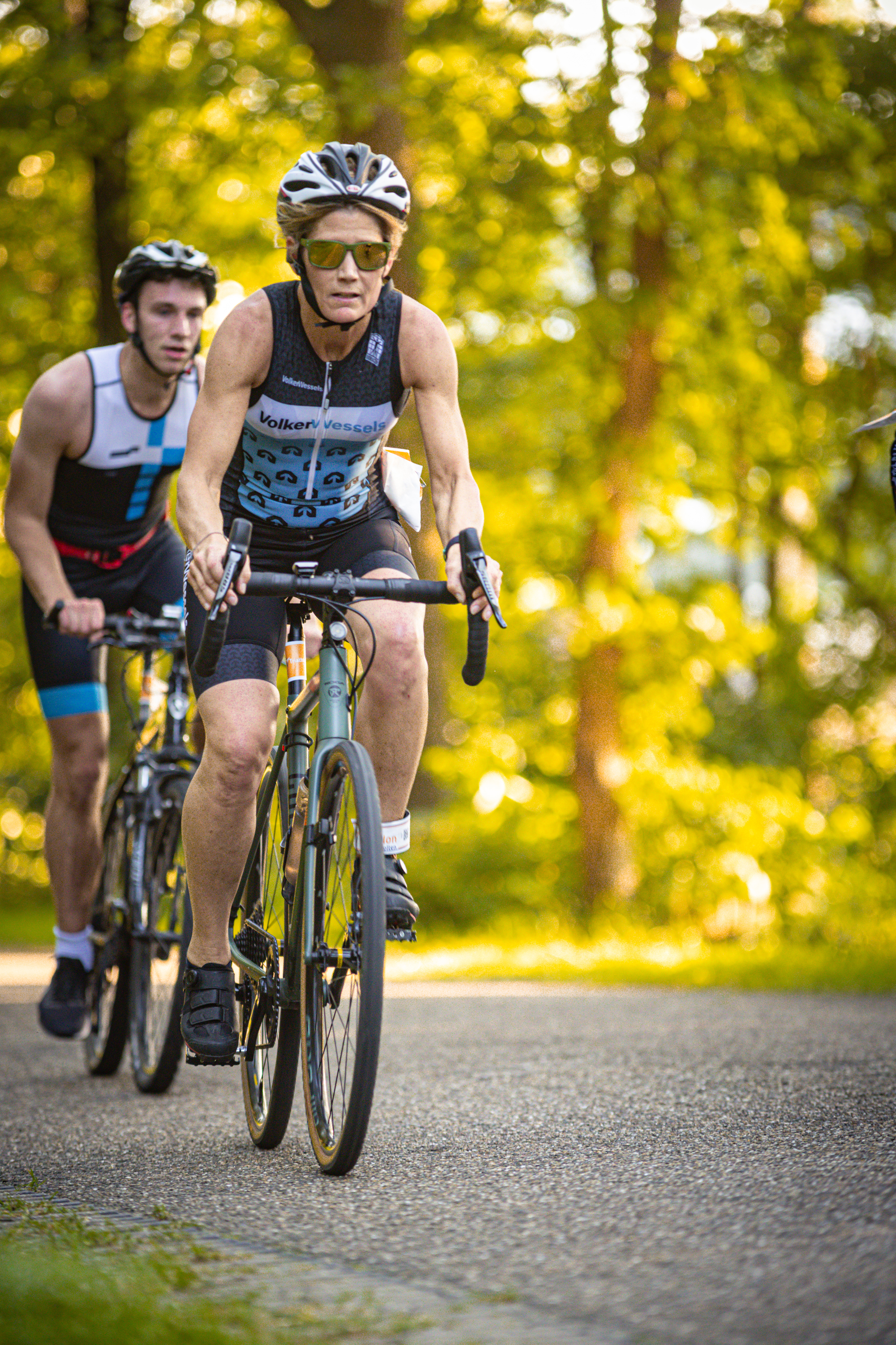 Two men are riding bikes on a road. One man is wearing a shirt that says "Holten Triathlon".