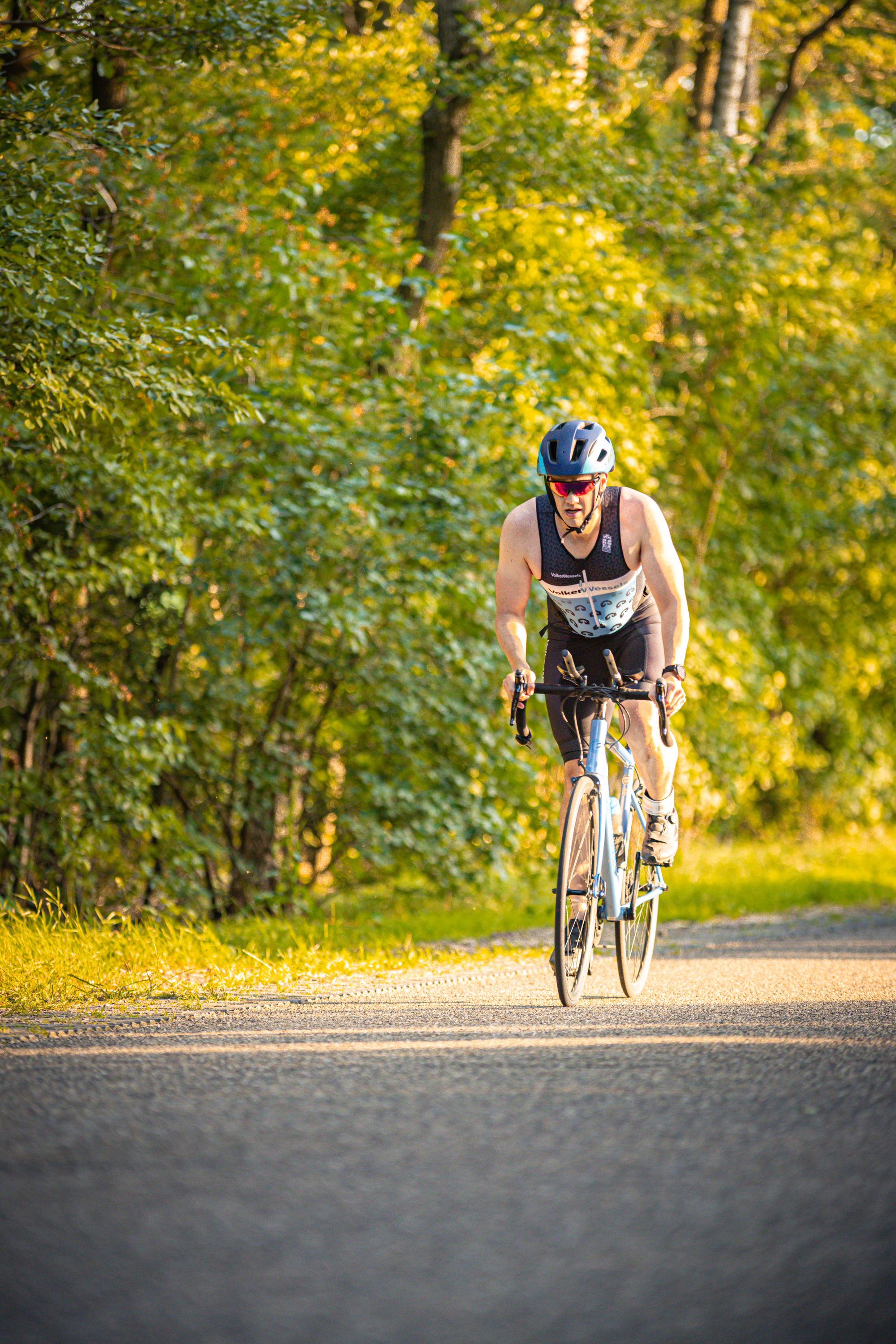 A triathlete is biking through the woods, wearing a black tank top and bike helmet.