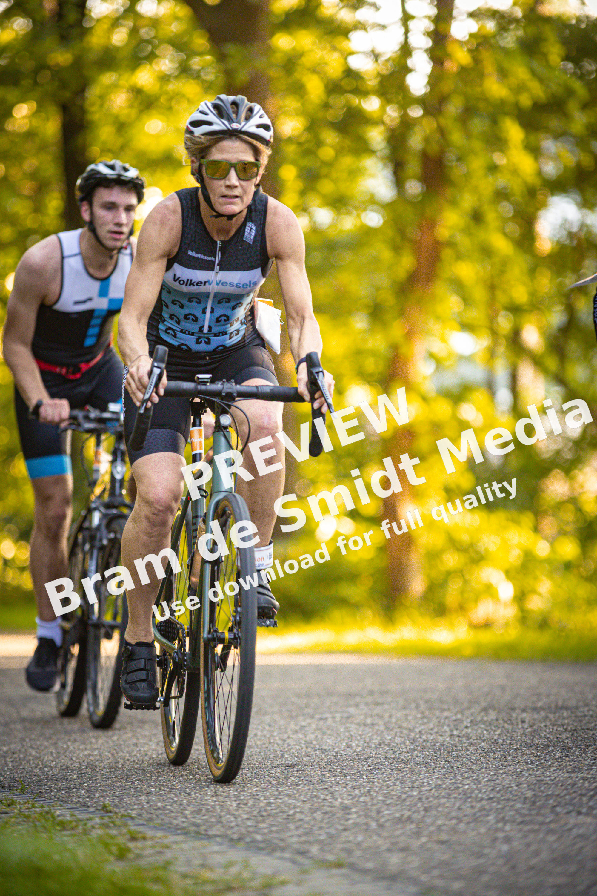 Two men are riding bikes on a road. One man is wearing a shirt that says "Holten Triathlon".