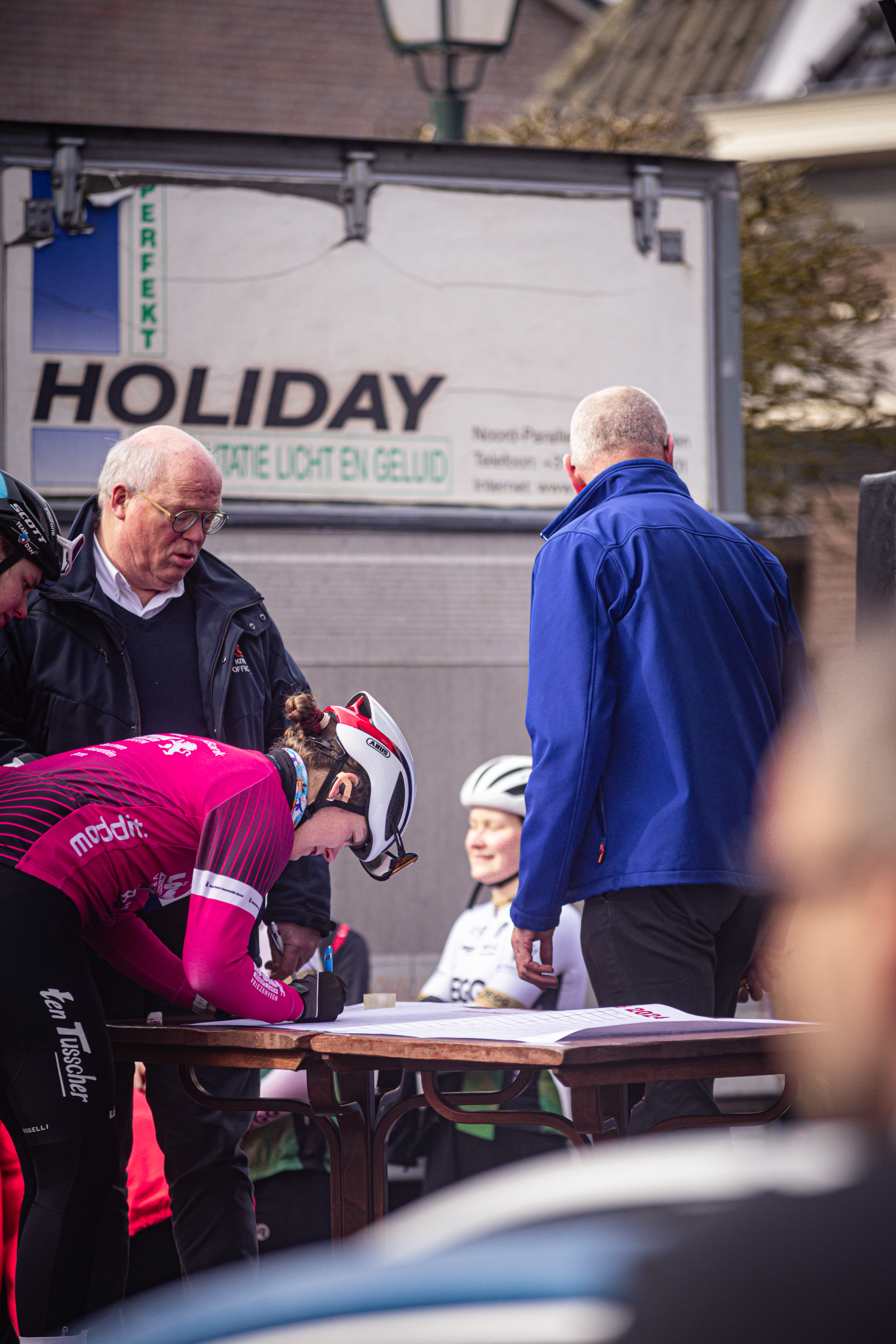 Three cyclists stand in front of a sign for Holiday.