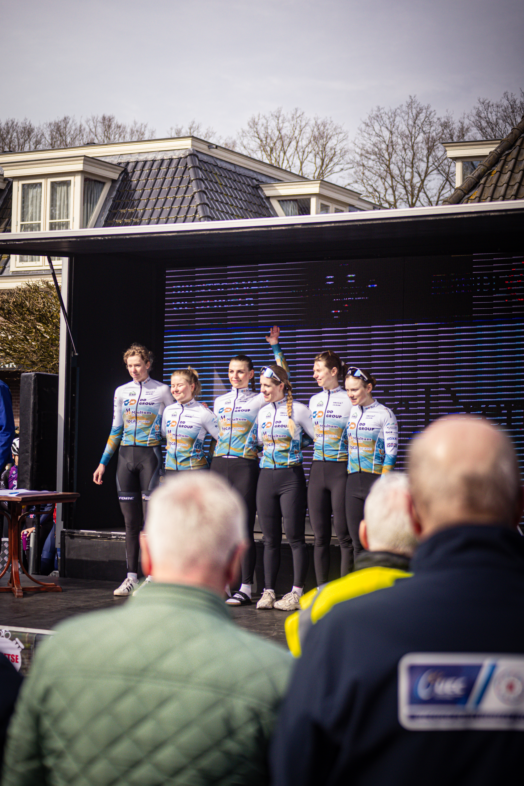 A group of cyclists with the words "Drentse 8 van Westerveld" on their jerseys, are standing together in front of a building.