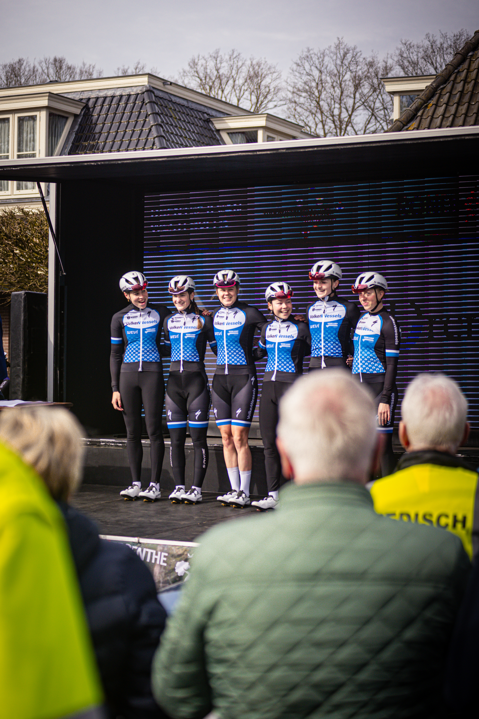 A group of cyclists stand on a stage in front of a large screen, with spectators looking on.