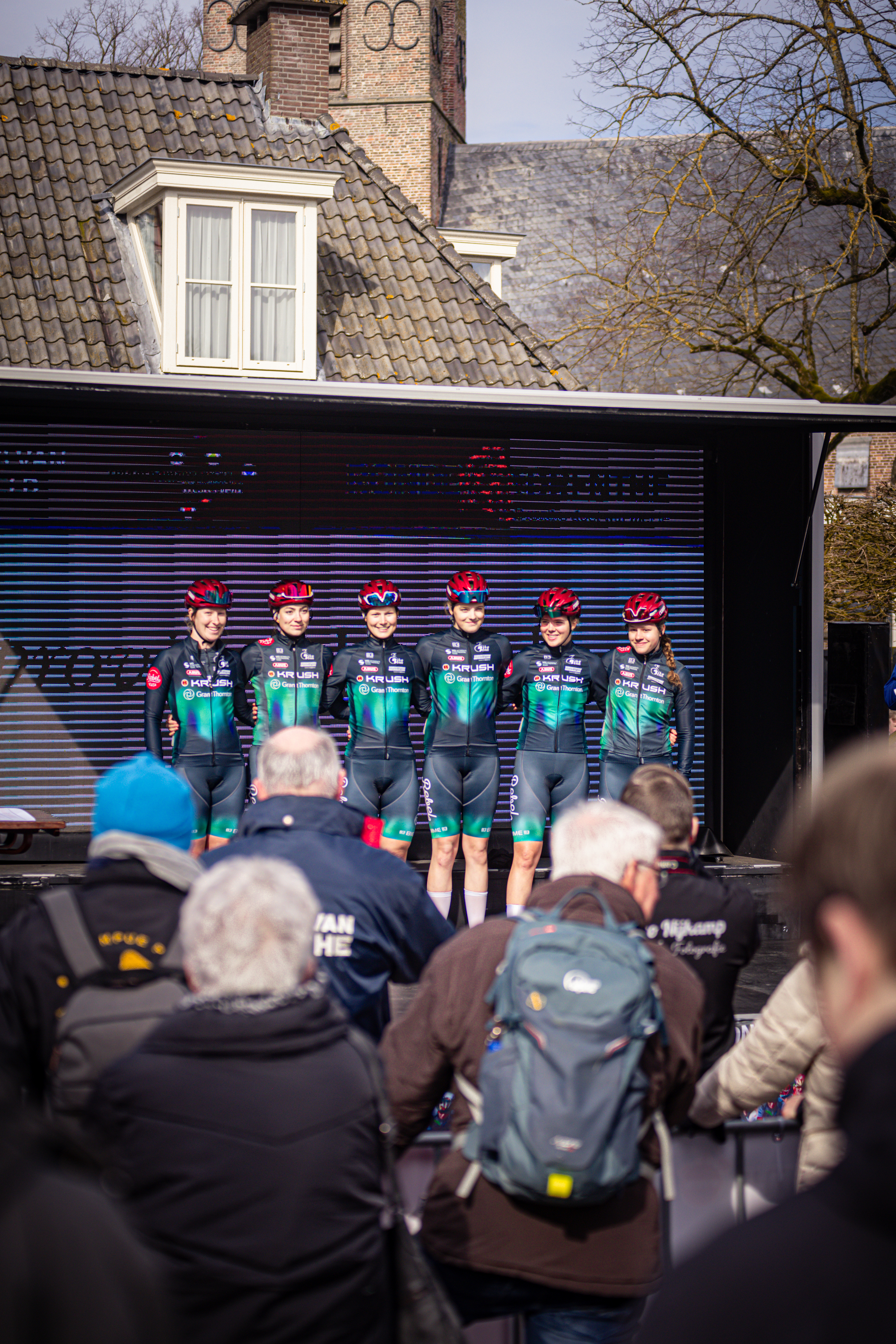 Eight cyclists standing together in front of a building and trees, wearing red helmets.