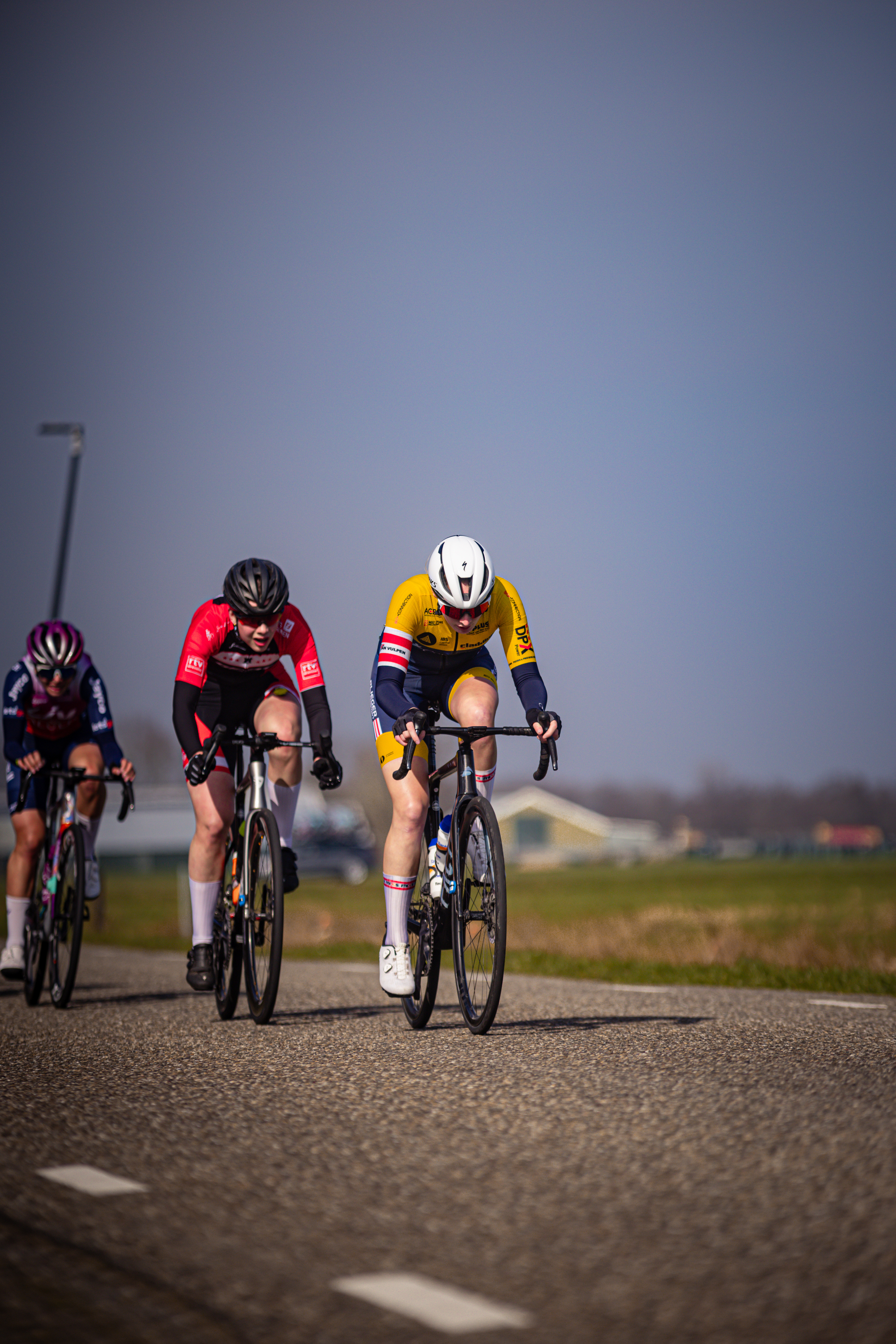 Three cyclists are on a road and two of them are wearing red shirts.