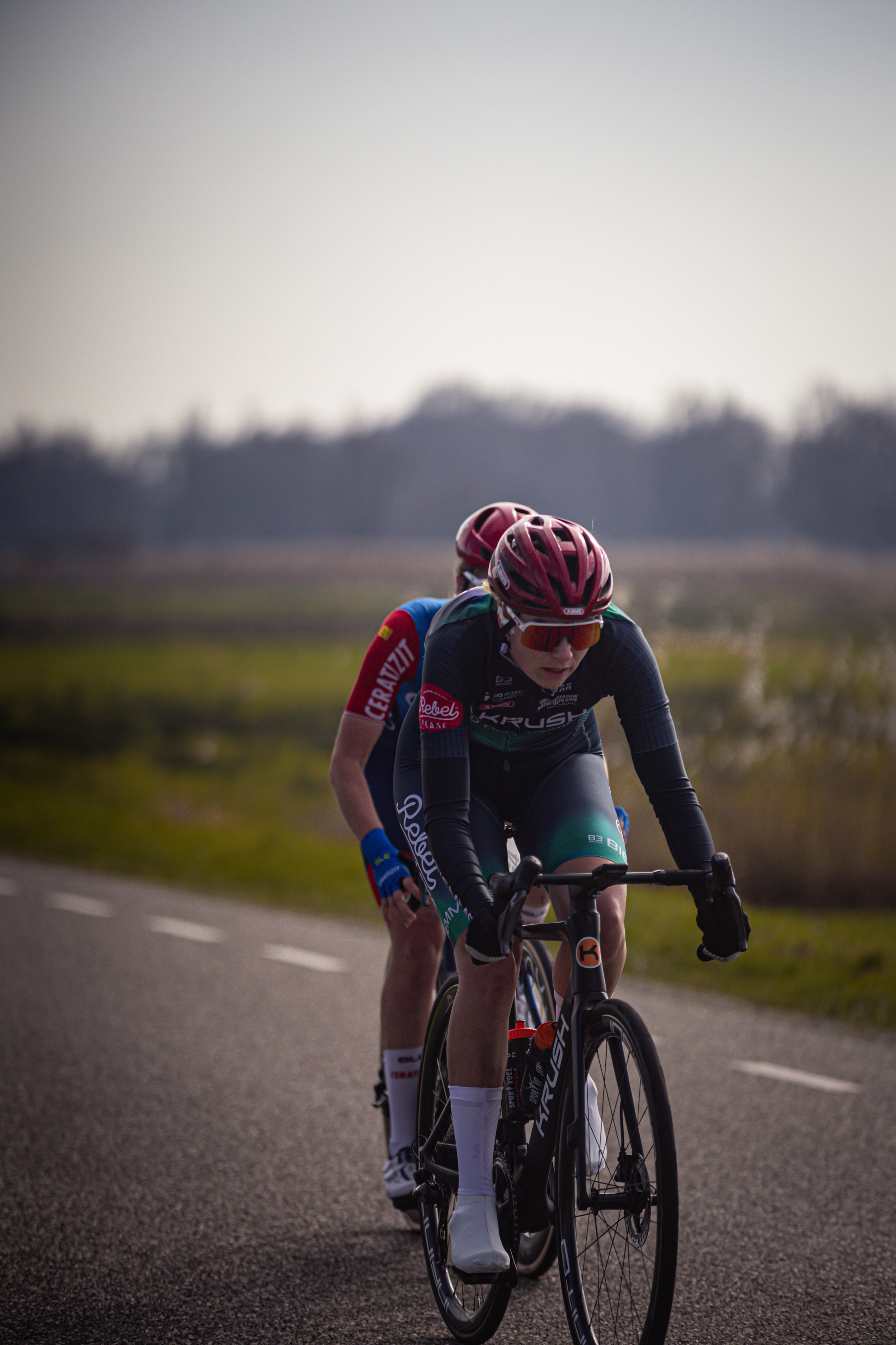 Two people on bikes going along a road with Drentse 8 van Westerveld in the background.