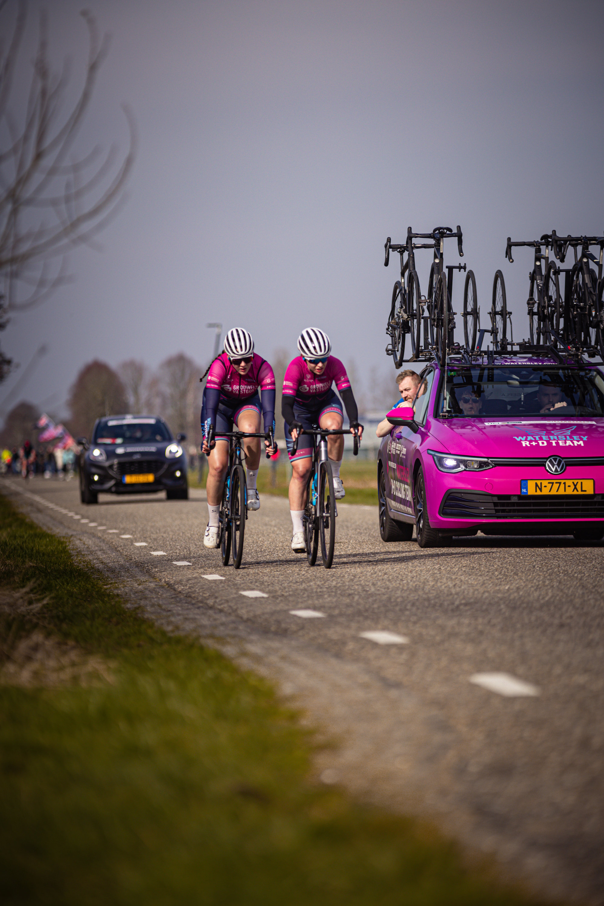 Three cyclists racing on a road near pink car with the word "Wielrennen" written on the top.