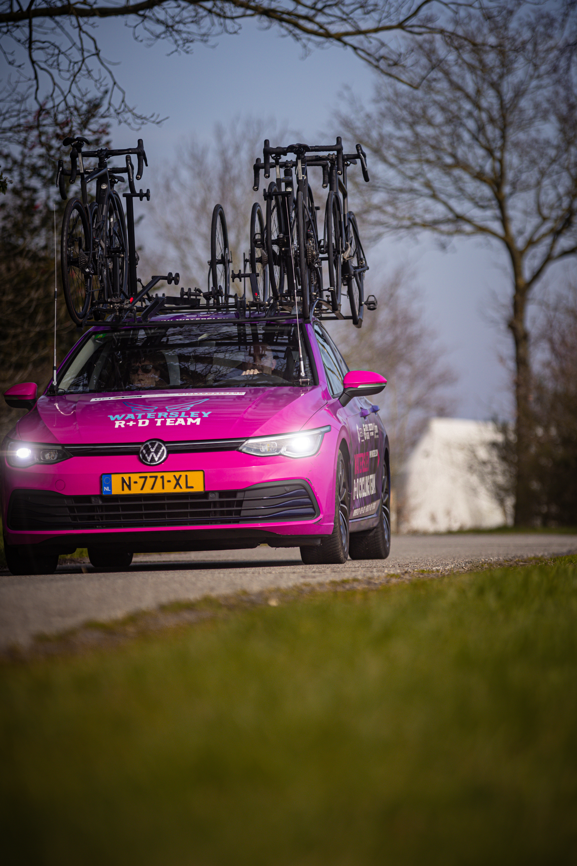 A pink Volkswagen Golf GTI car. The car has a bike rack on the front and is parked on a street near trees.