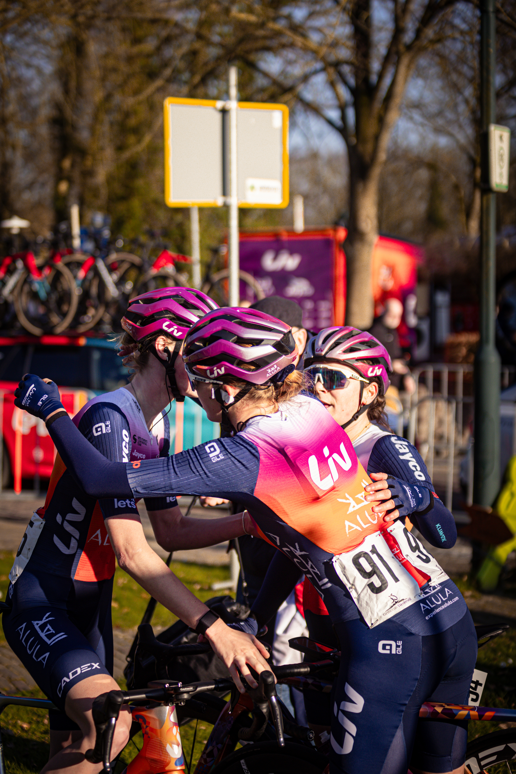 A pair of cyclists wearing pink helmets are embracing with one another.