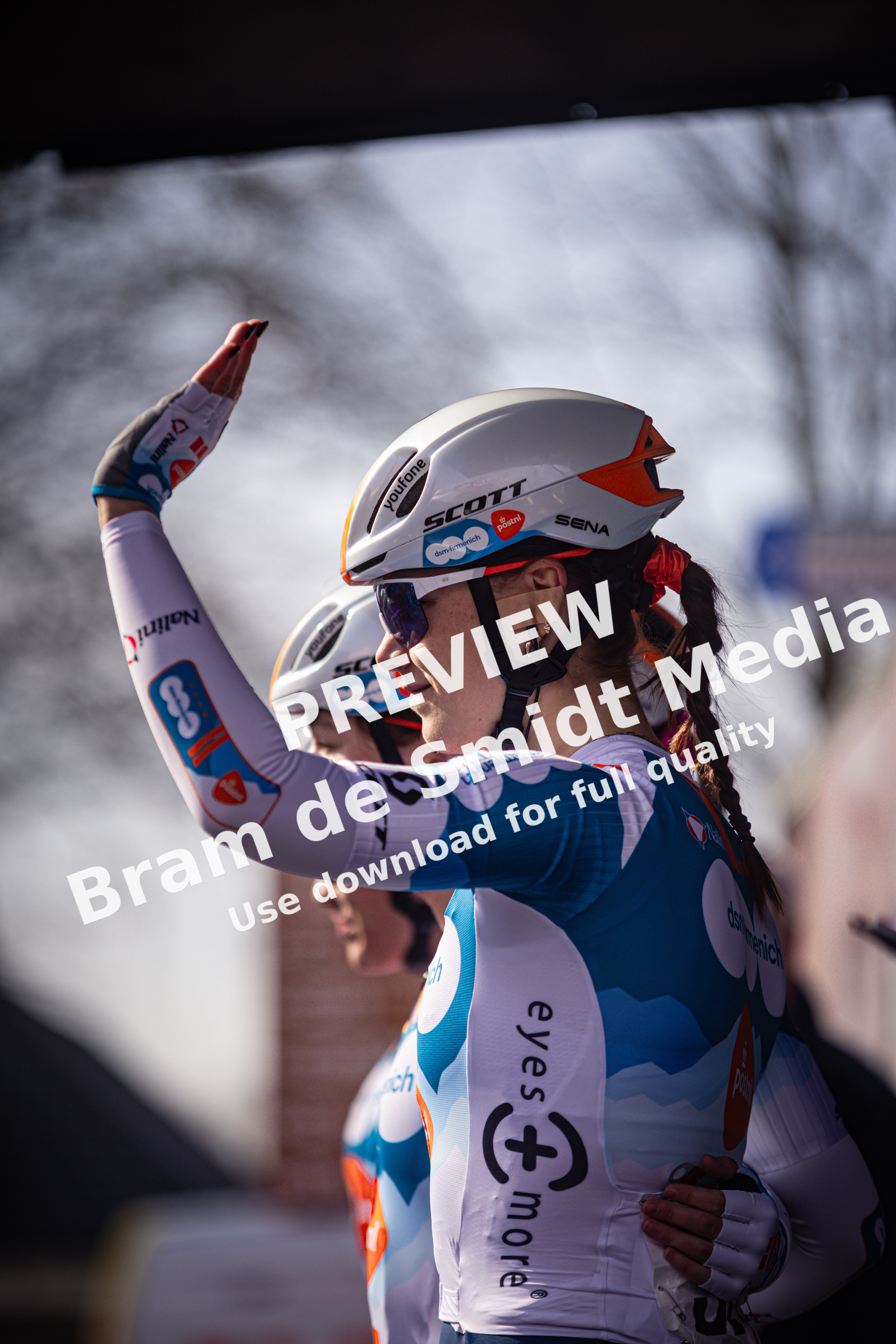 A cyclist with her arm raised, wearing a helmet and uniform for the Wielrennen race.