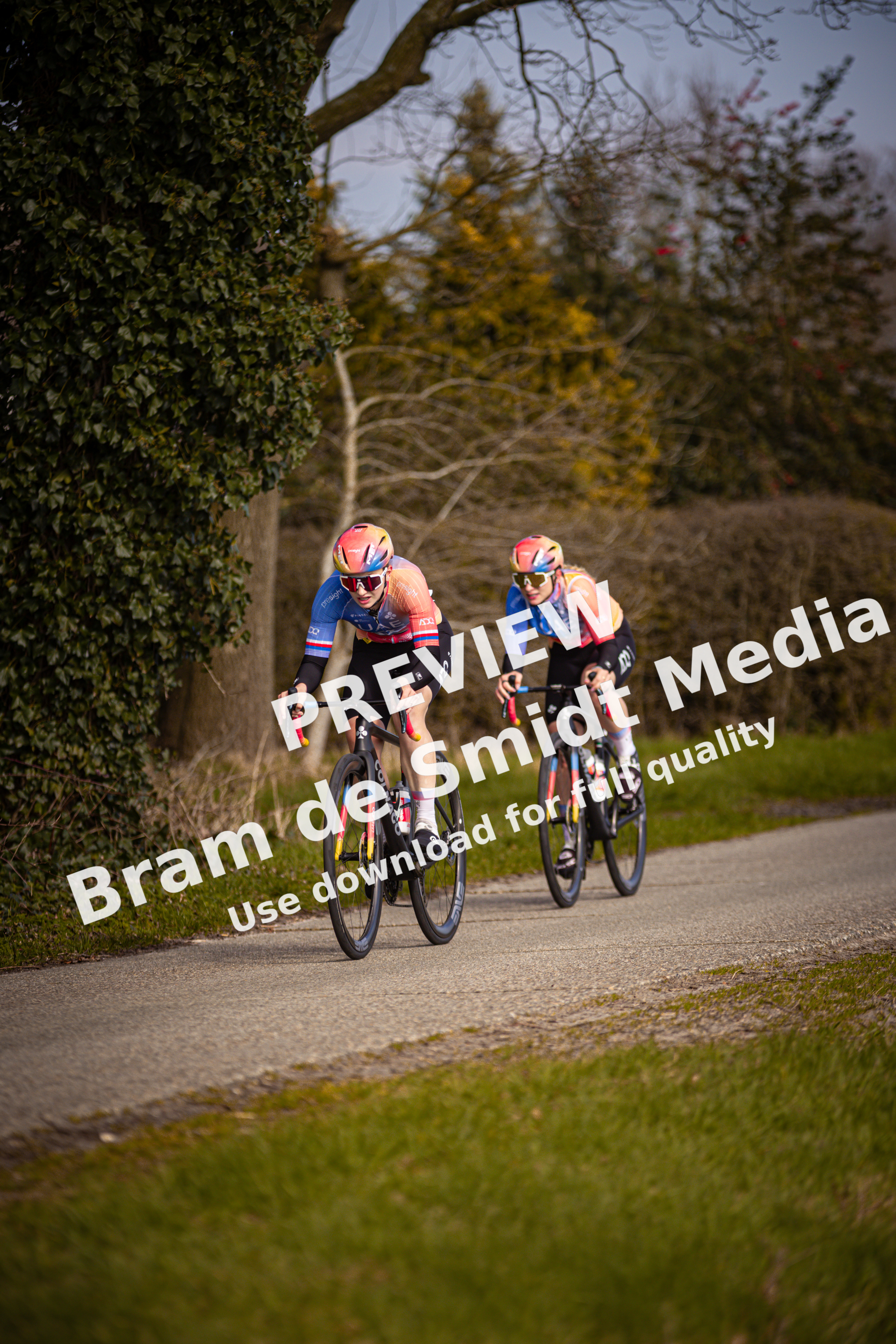 Two cyclists with helmets of different colors are wearing matching team colors and are riding their bikes down a narrow road.